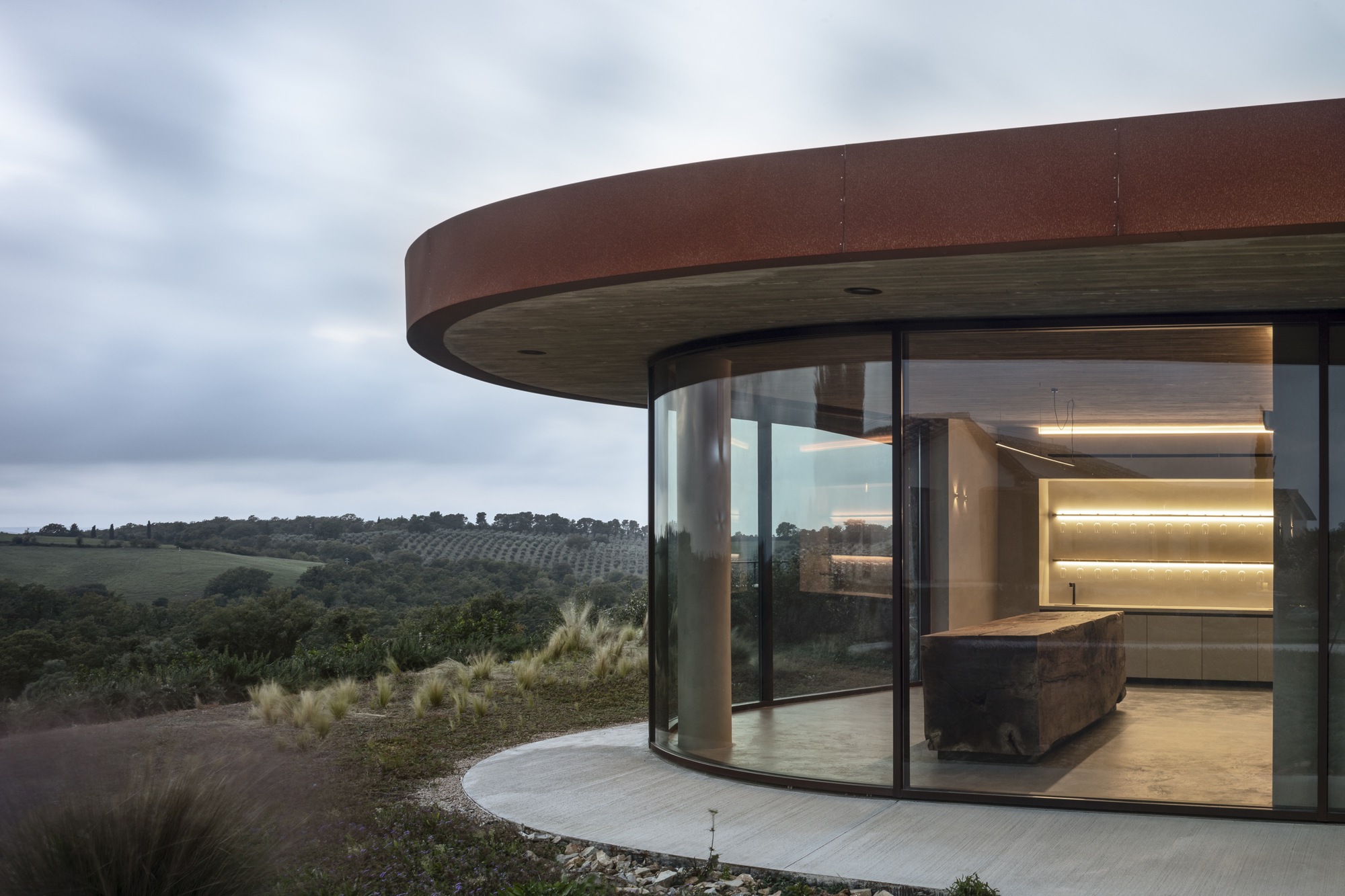 Curved glass walls beneath a corten steel overhang opening to a terrace with views of distant orchards