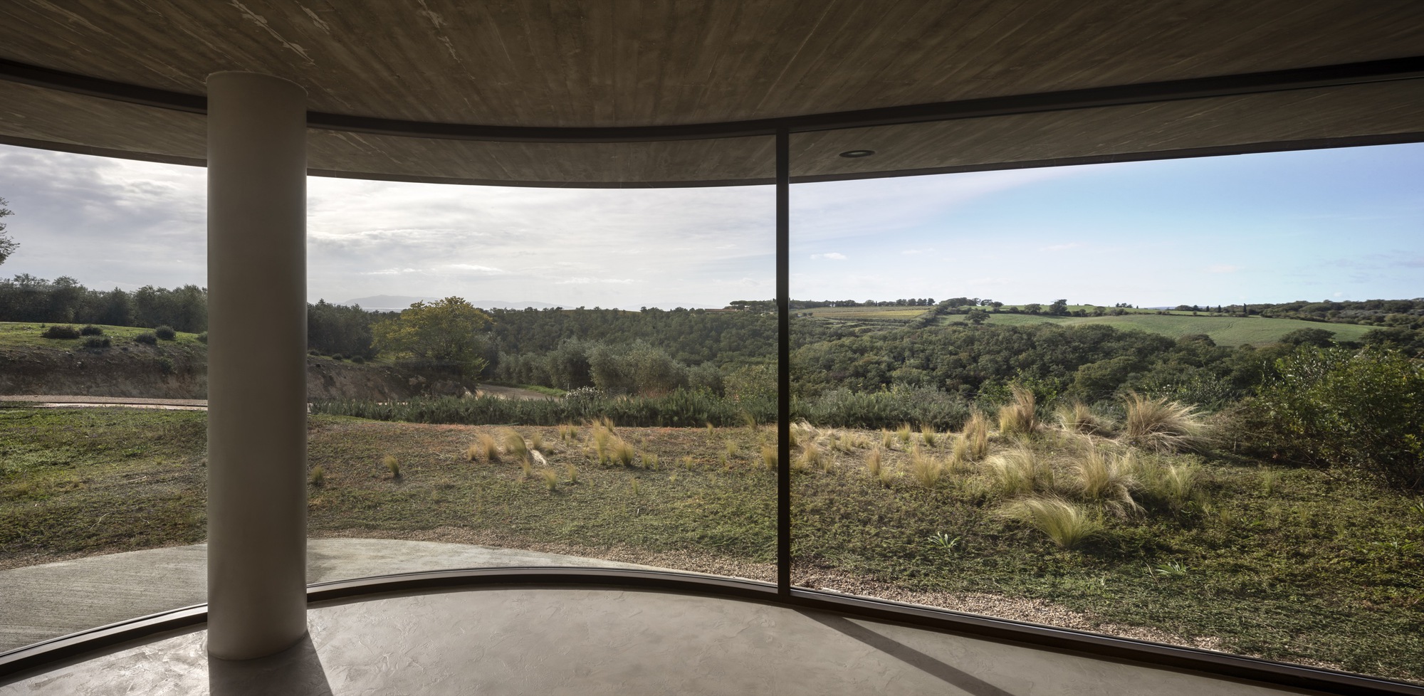 Curved floor-to-ceiling glazing framing olive groves with concrete floor and cylindrical columns under cloudy skies