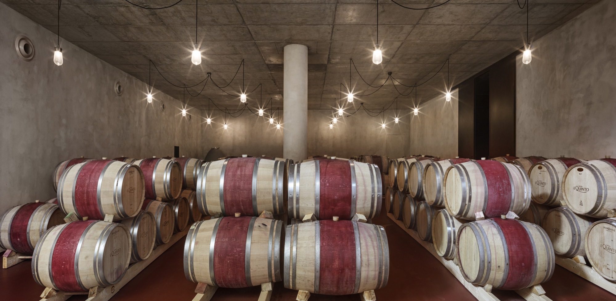 Barrel room with exposed concrete ceiling and string lights illuminating stacked wine barrels