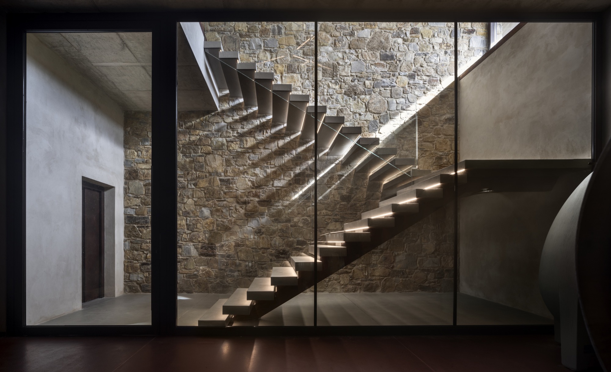 Floating steel staircase against an exposed stone wall illuminated by natural light from above