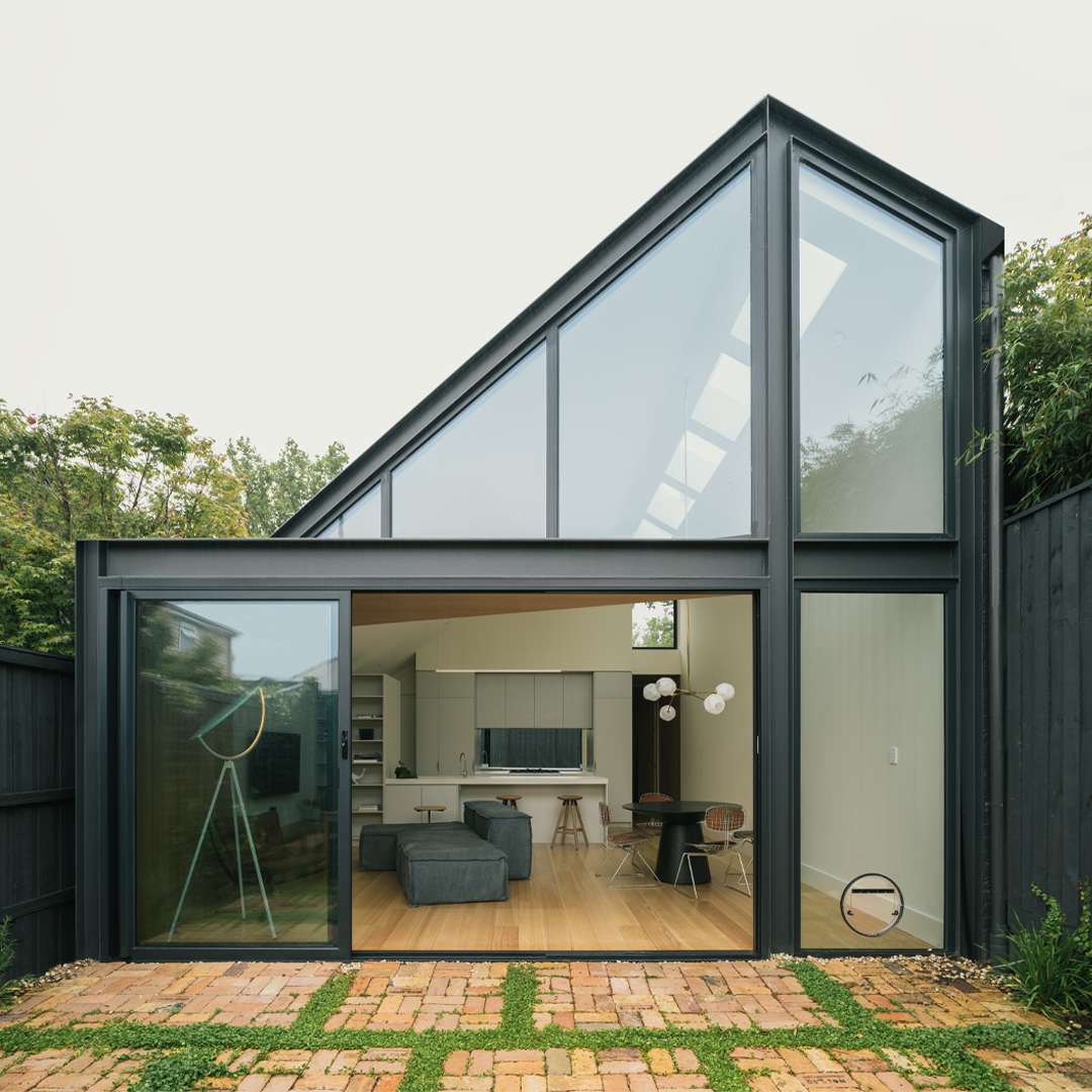 Rear garden elevation with angled glazing and black steel frames opening onto a brick and grass courtyard