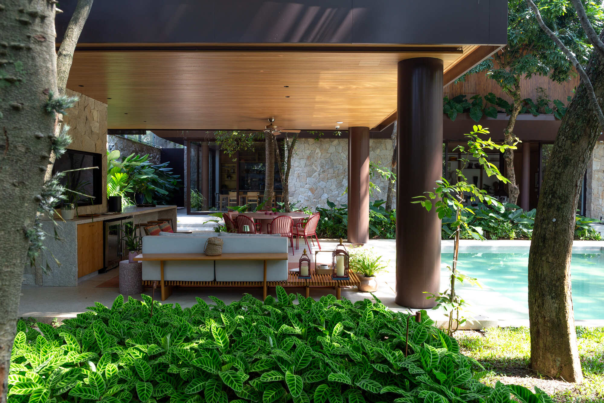 Open living pavilion with timber ceiling and cylindrical column opening to pool and garden through existing trees