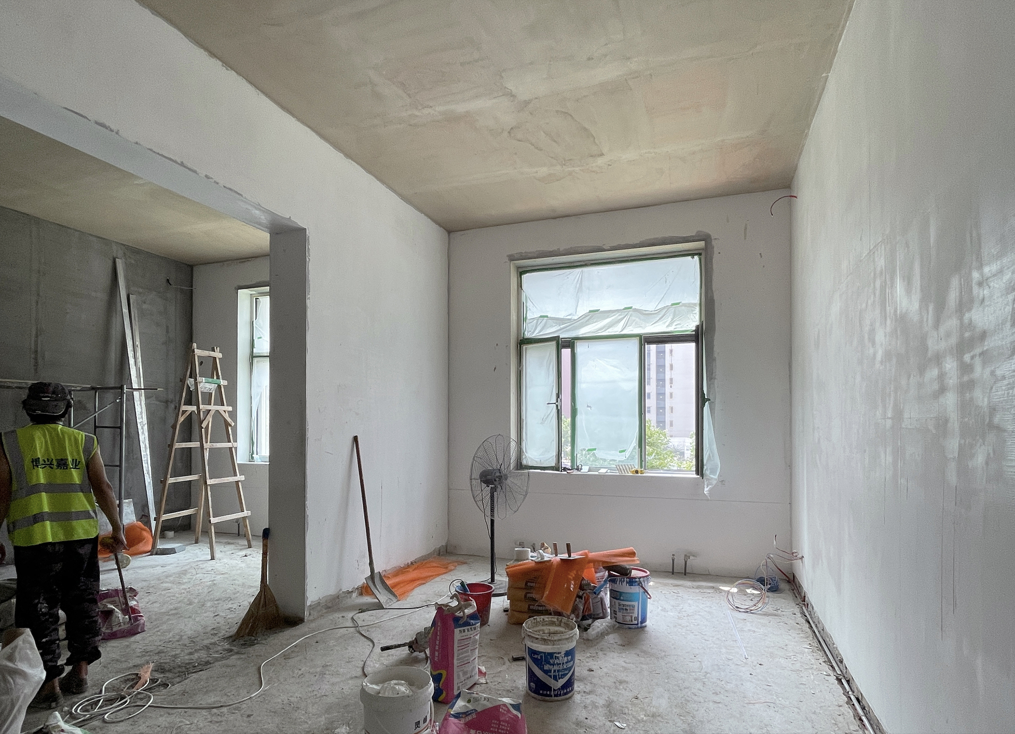 Plastered room under construction with a worker in high-visibility vest and paint buckets near the window
