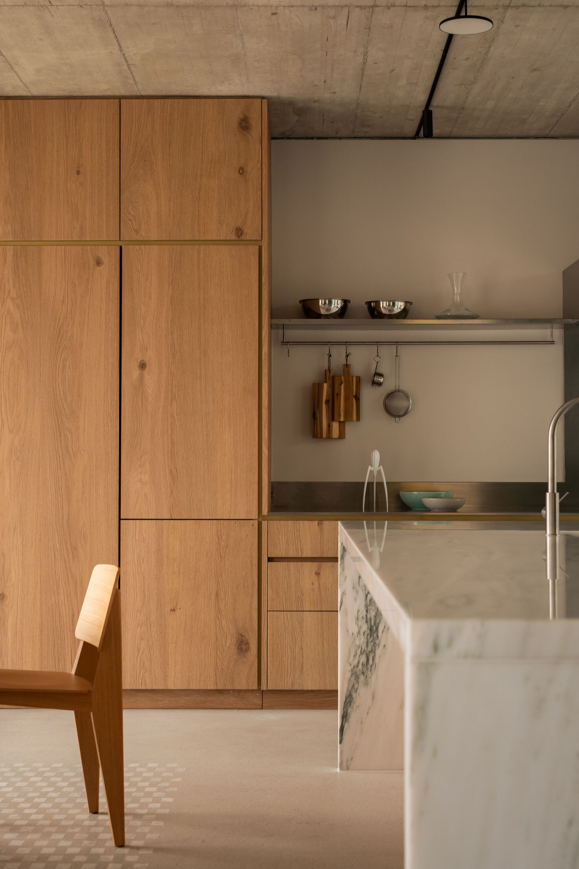 Kitchen corner with oak paneled cabinetry and marble island under exposed concrete ceiling with hanging utensils