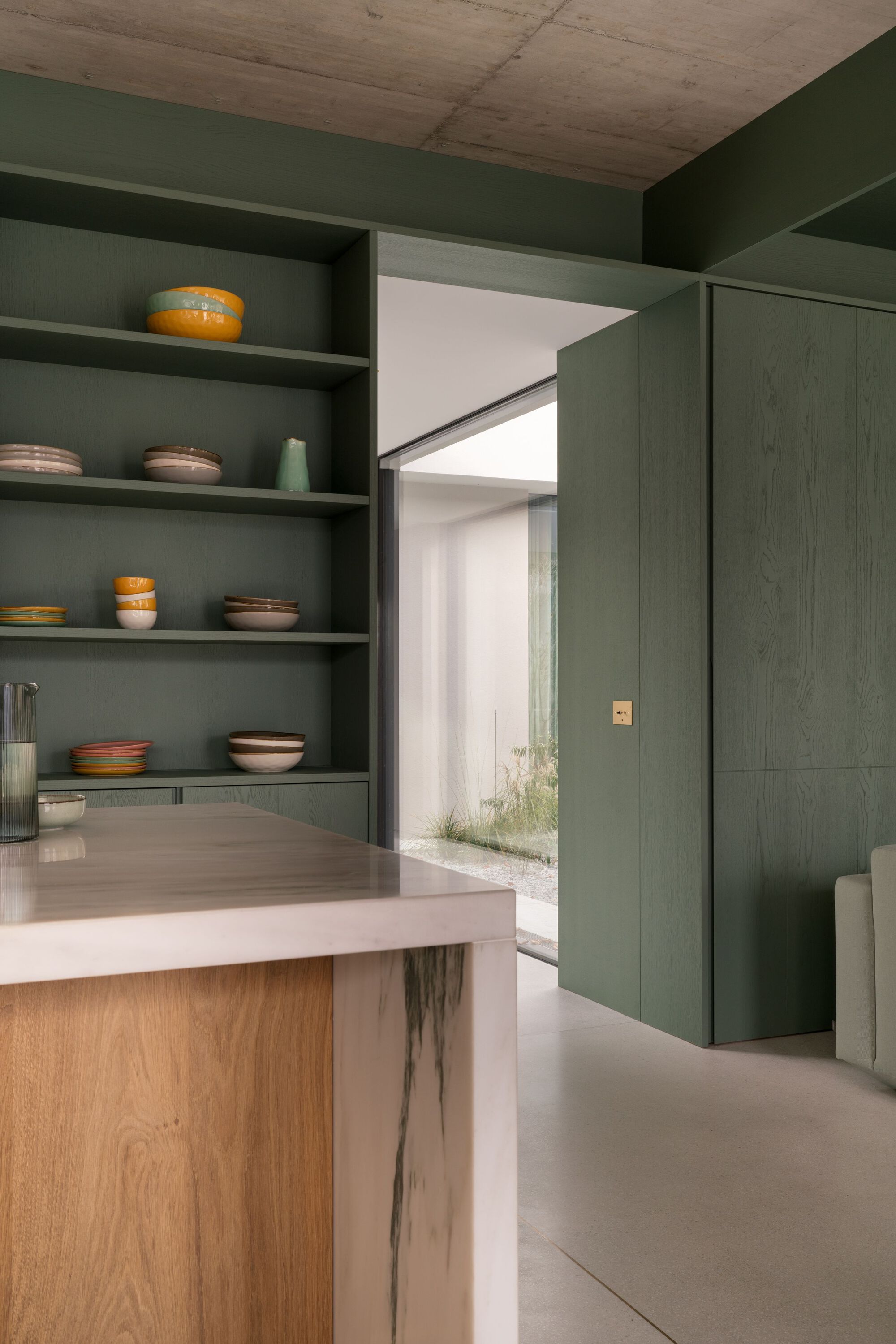 Kitchen island with green shelving displaying ceramic bowls and view through to courtyard with planted grasses