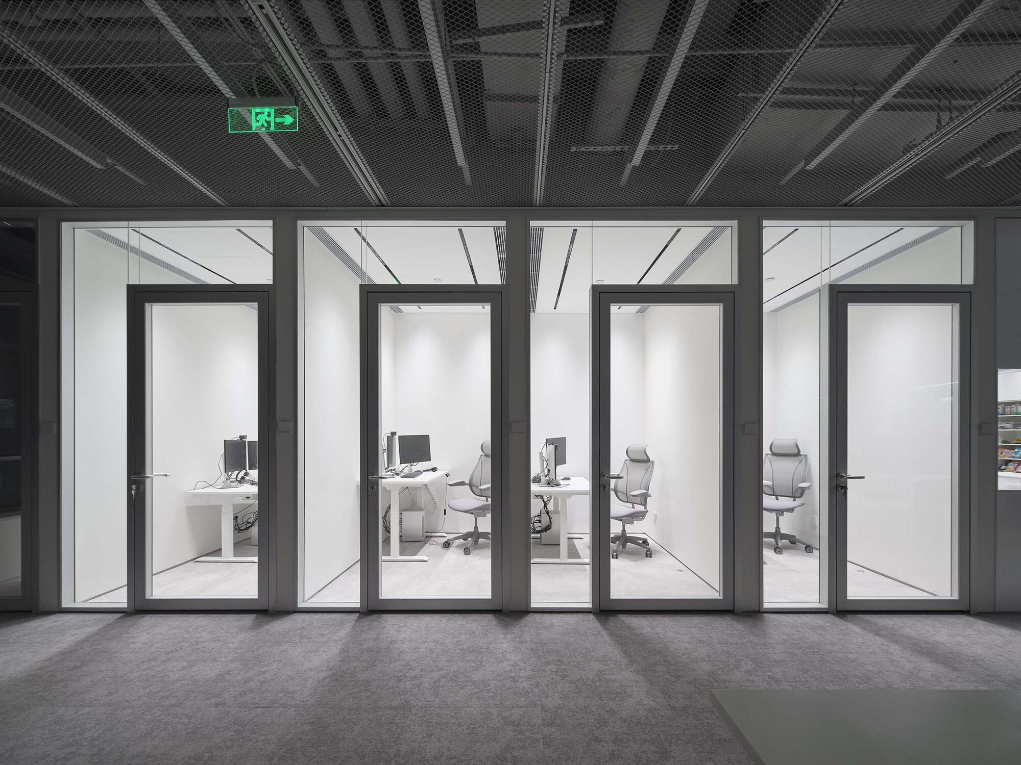 Four glass-walled office pods viewed from a corridor with exposed black ceiling and emergency exit sign