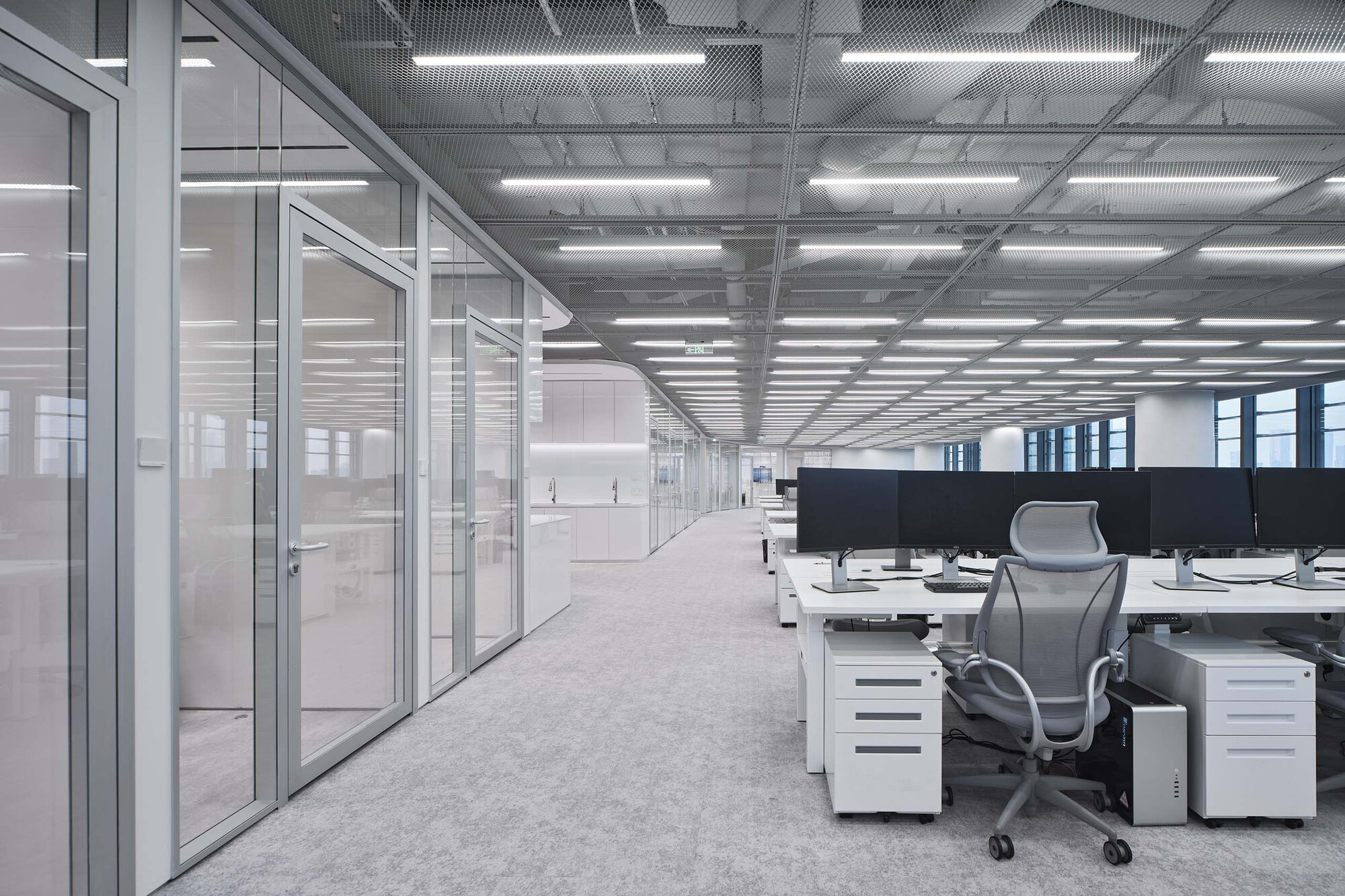 Workstation area with exposed ceiling ducts and rows of desks beside enclosed glass offices