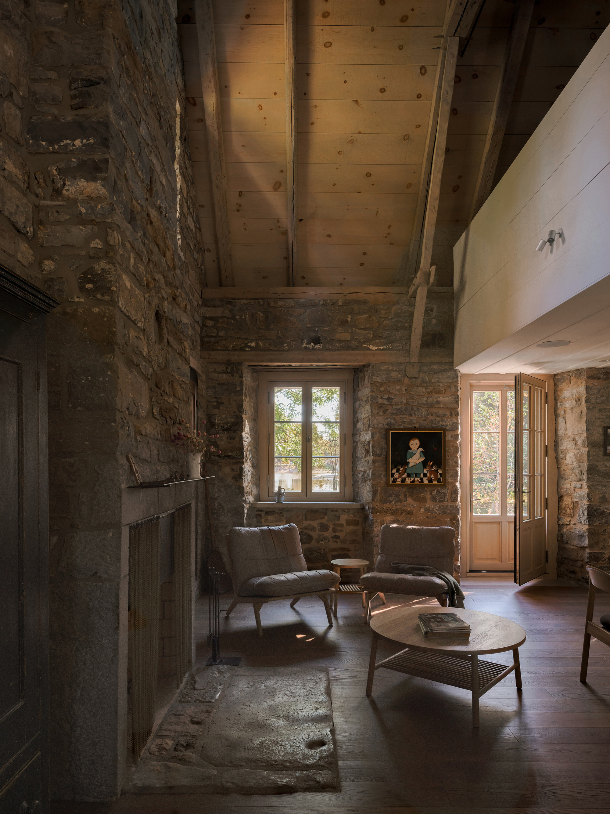 Double-height living room with exposed stone walls, timber ceiling beams and afternoon light through casement windows