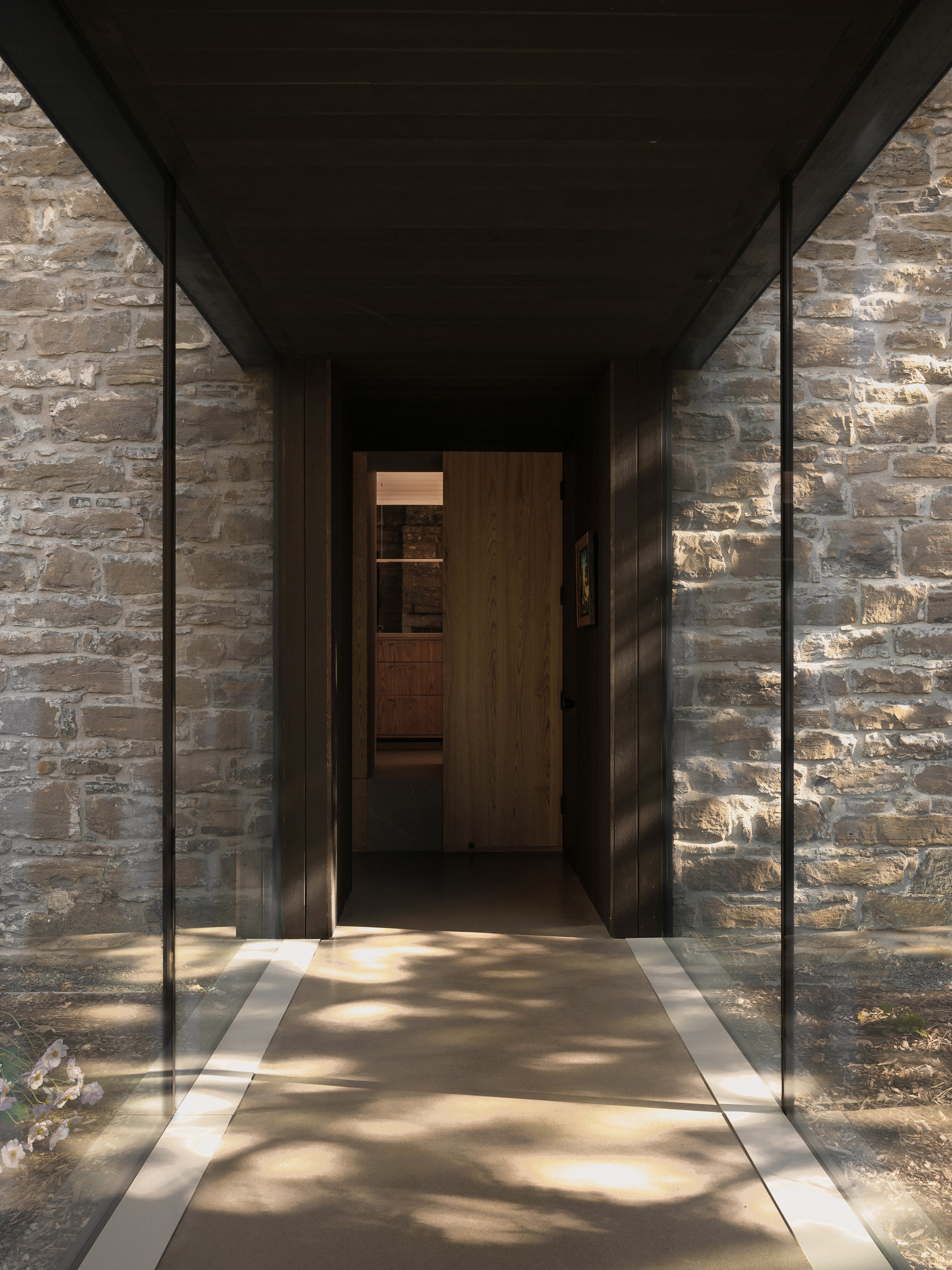 Covered entry passage with steel-framed glazing flanking timber doors and stone walls in dappled sunlight