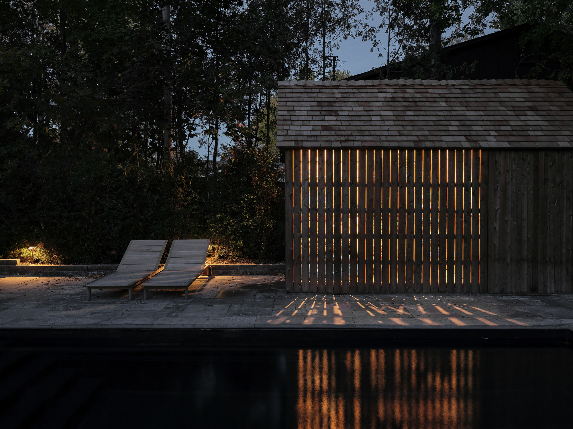 Vertical timber screen with interior light casting striped shadows on the pool deck at dusk