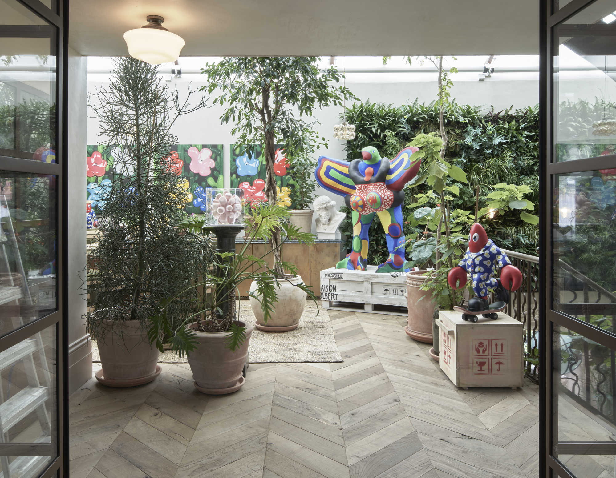 Interior courtyard with herringbone wood floor, potted plants, and colorful sculptures among steel-framed glazing