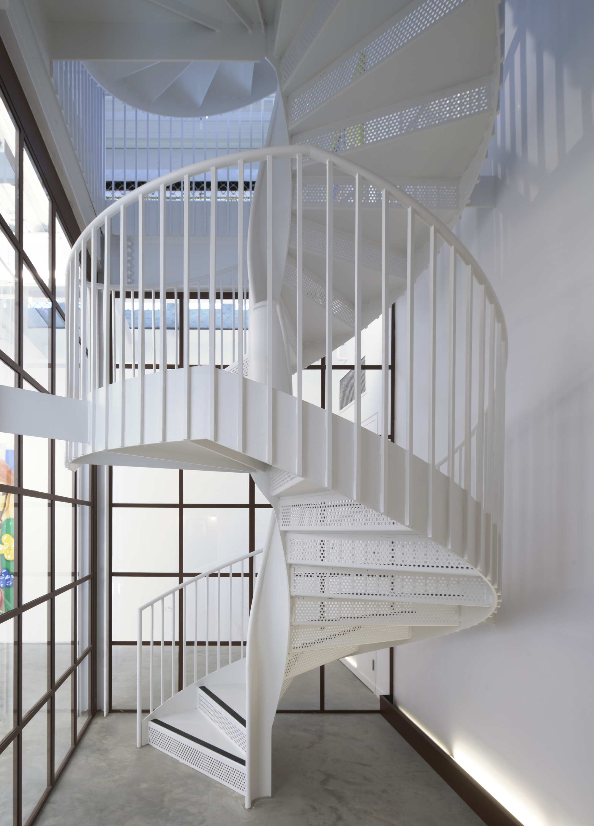 White spiral staircase with perforated metal treads and vertical bar railings beside steel-framed windows