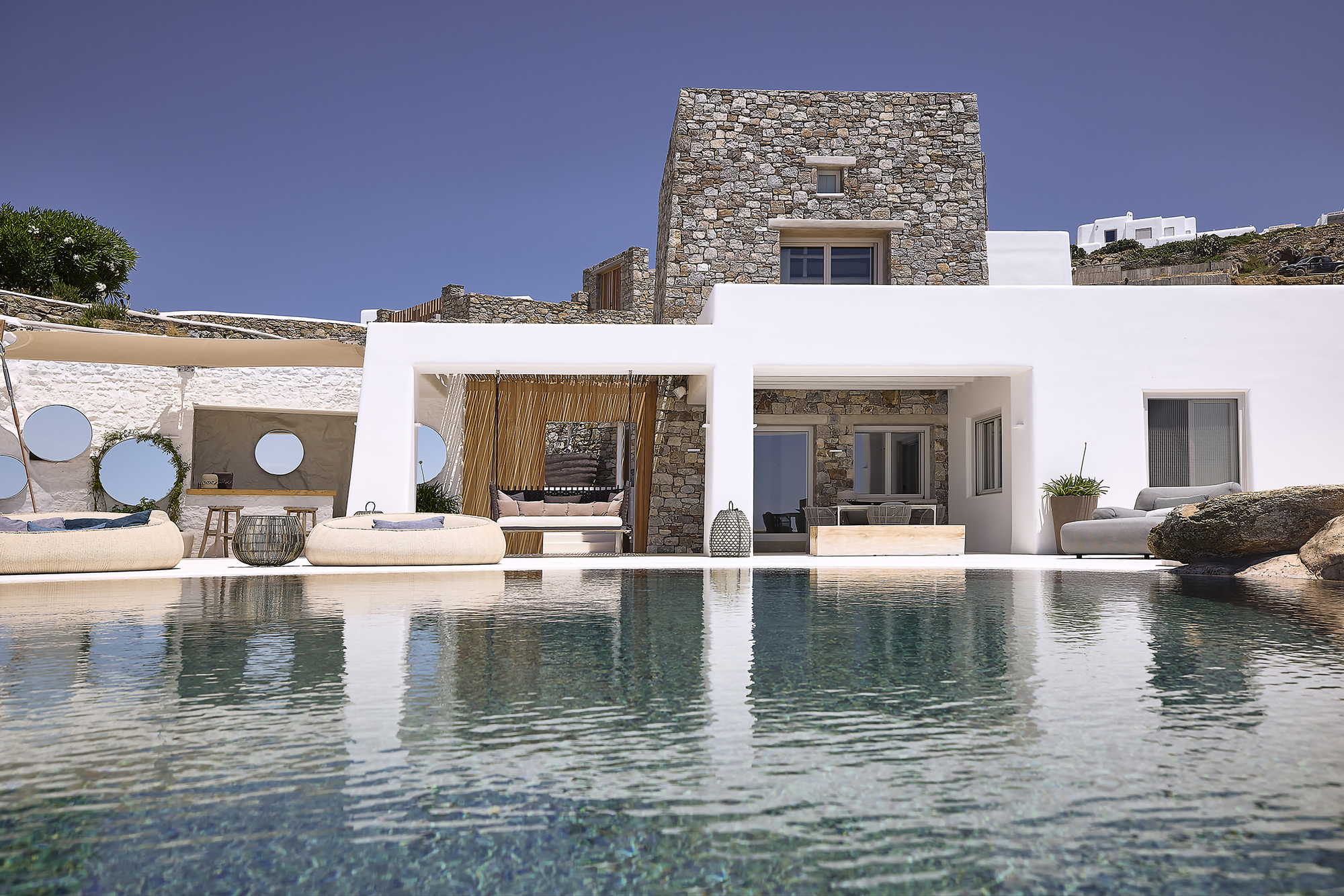 Pool deck with circular porthole screens in white pergola facing stone tower volume under clear sky