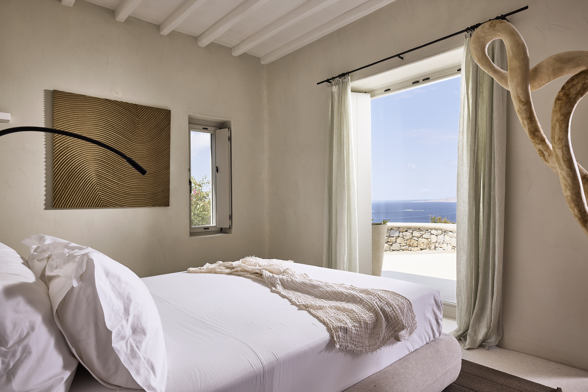 Bedroom window framing ocean view with stone wall terrace and linen curtains under blue sky