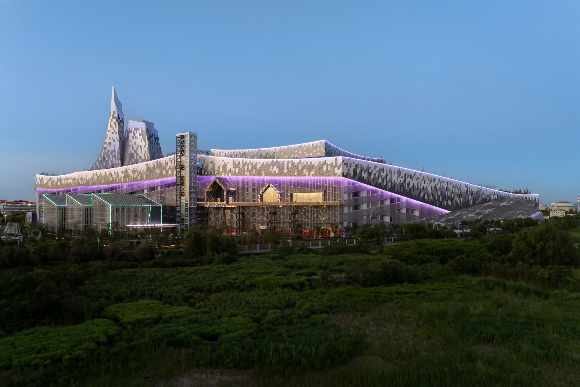 Illuminated perforated facade with purple accent lighting viewed across a green field at dusk