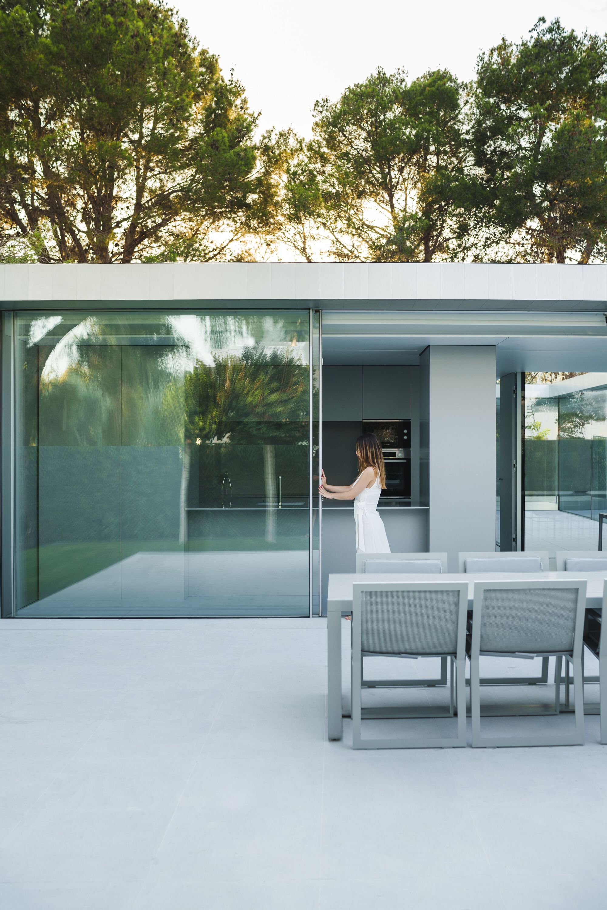 Outdoor dining terrace with glass sliding walls opening to kitchen and surrounding trees at dusk