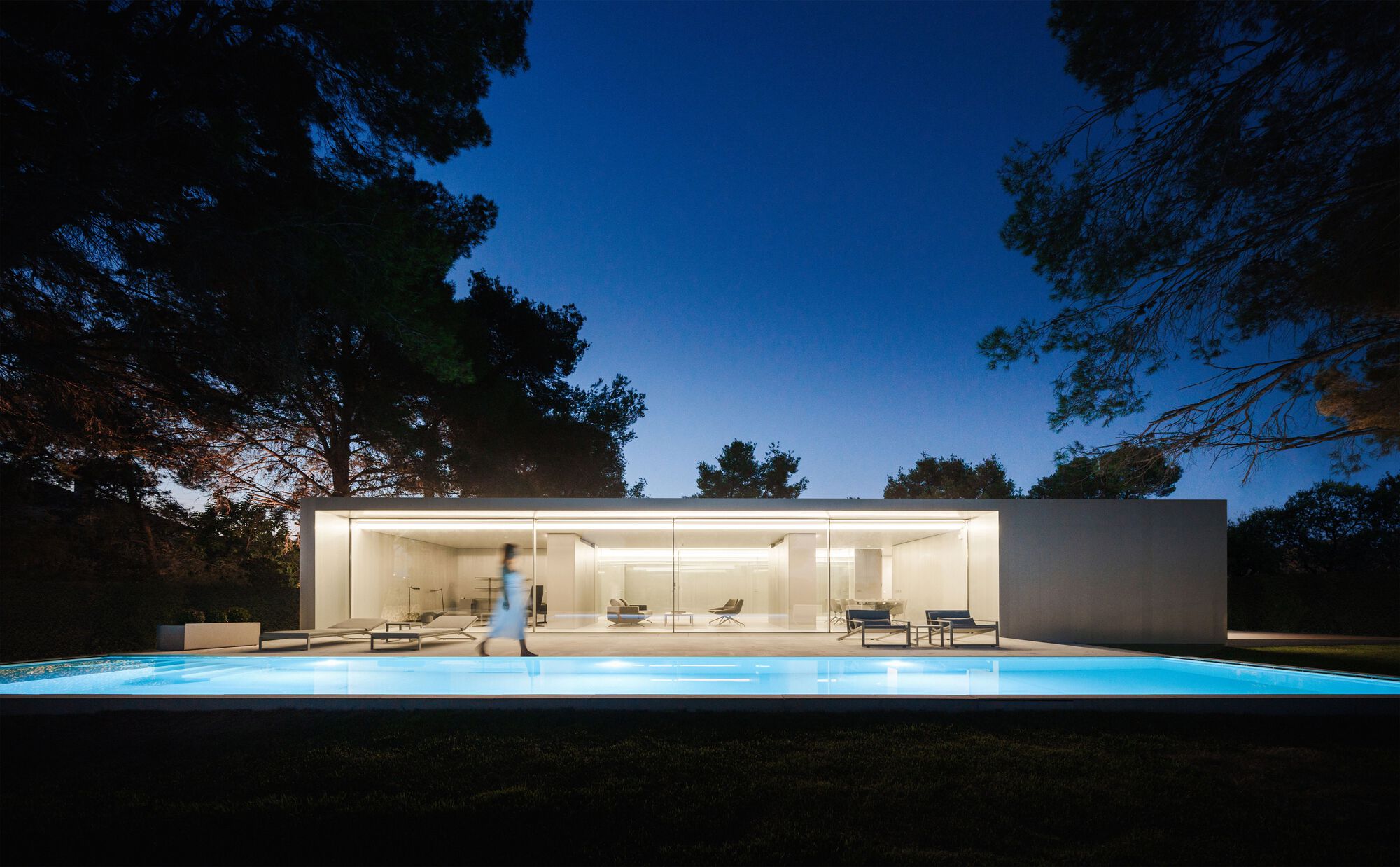 Illuminated glass facade and glowing pool terrace with a figure walking at twilight