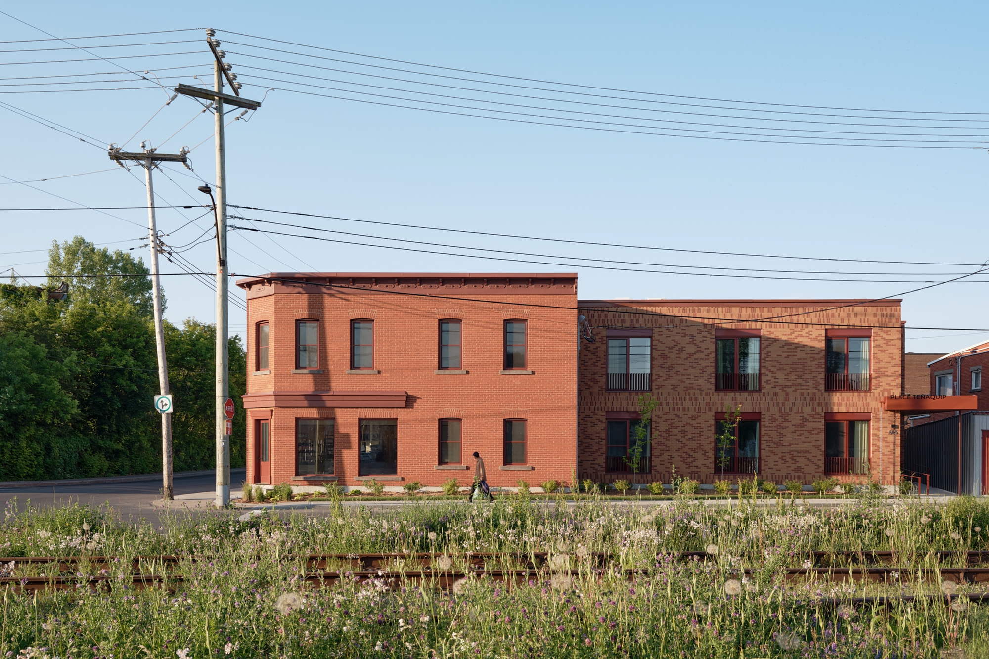 Street facade with varied brick textures across volumes and a person walking along the planted edge