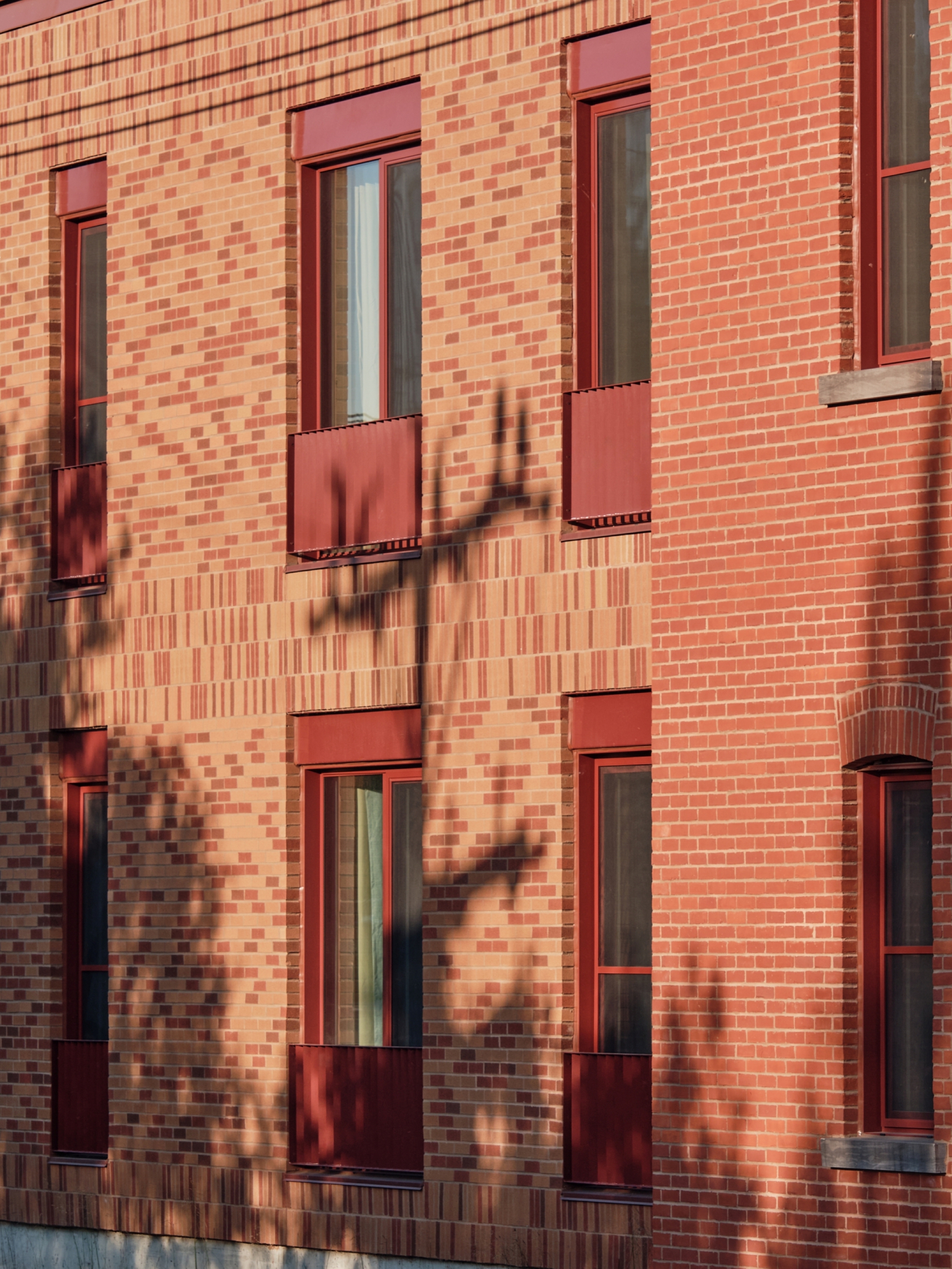 Facade detail showing patterned brickwork with tree shadows cast across windows and red metal panels