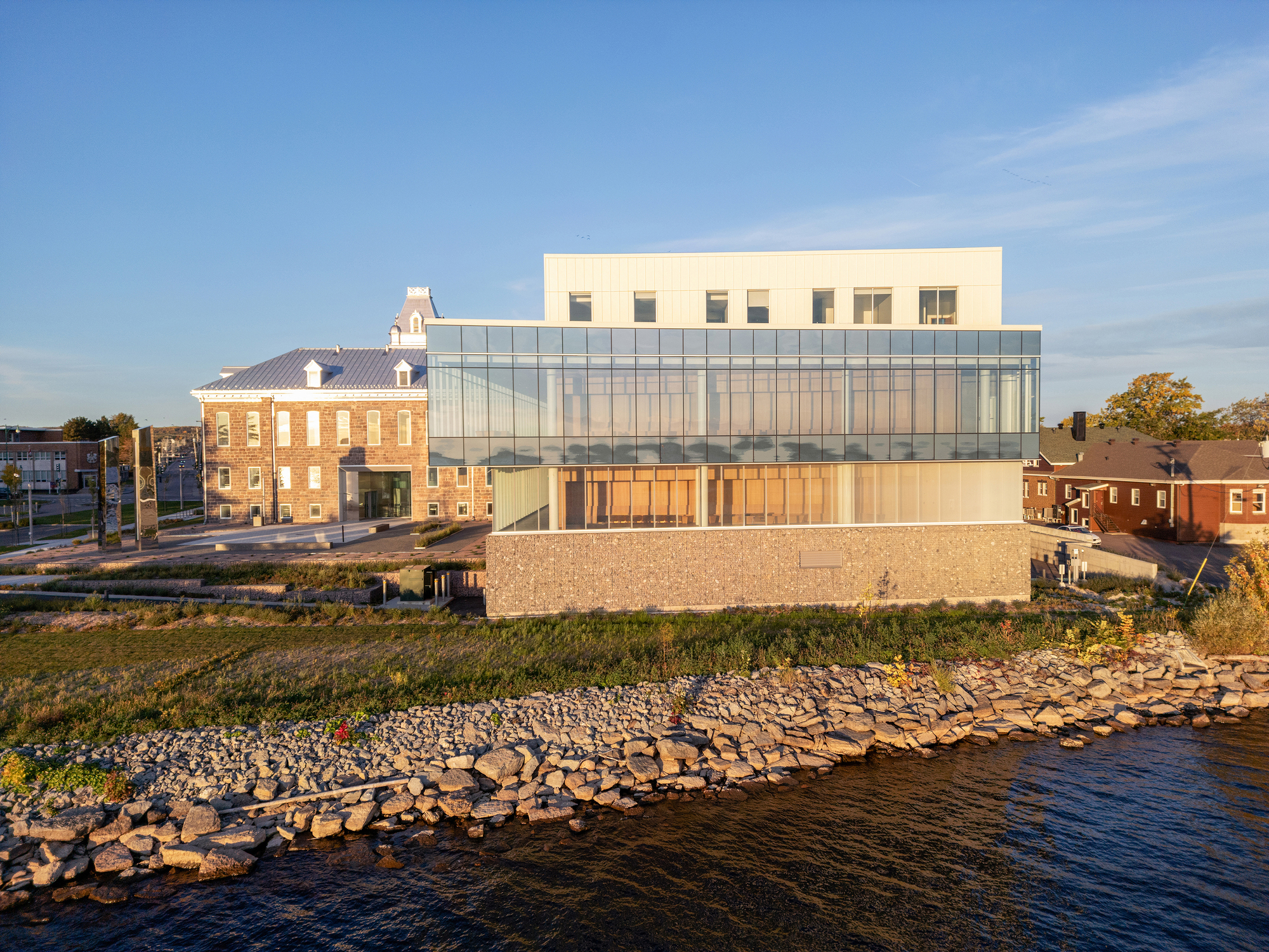 Waterfront elevation with glass curtain wall above a gabion stone base at golden hour