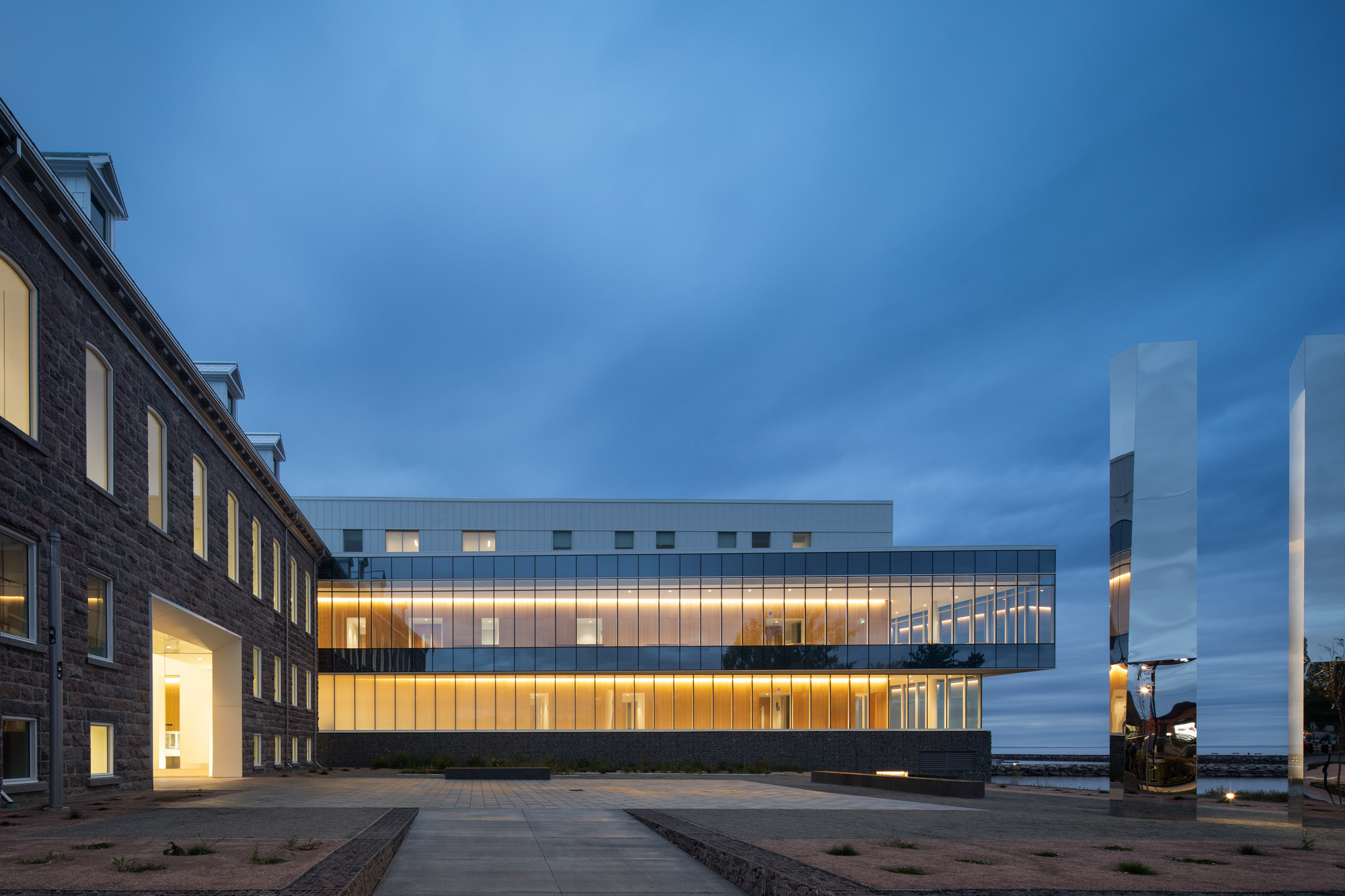 Illuminated glass facade extension alongside a brick heritage building at dusk