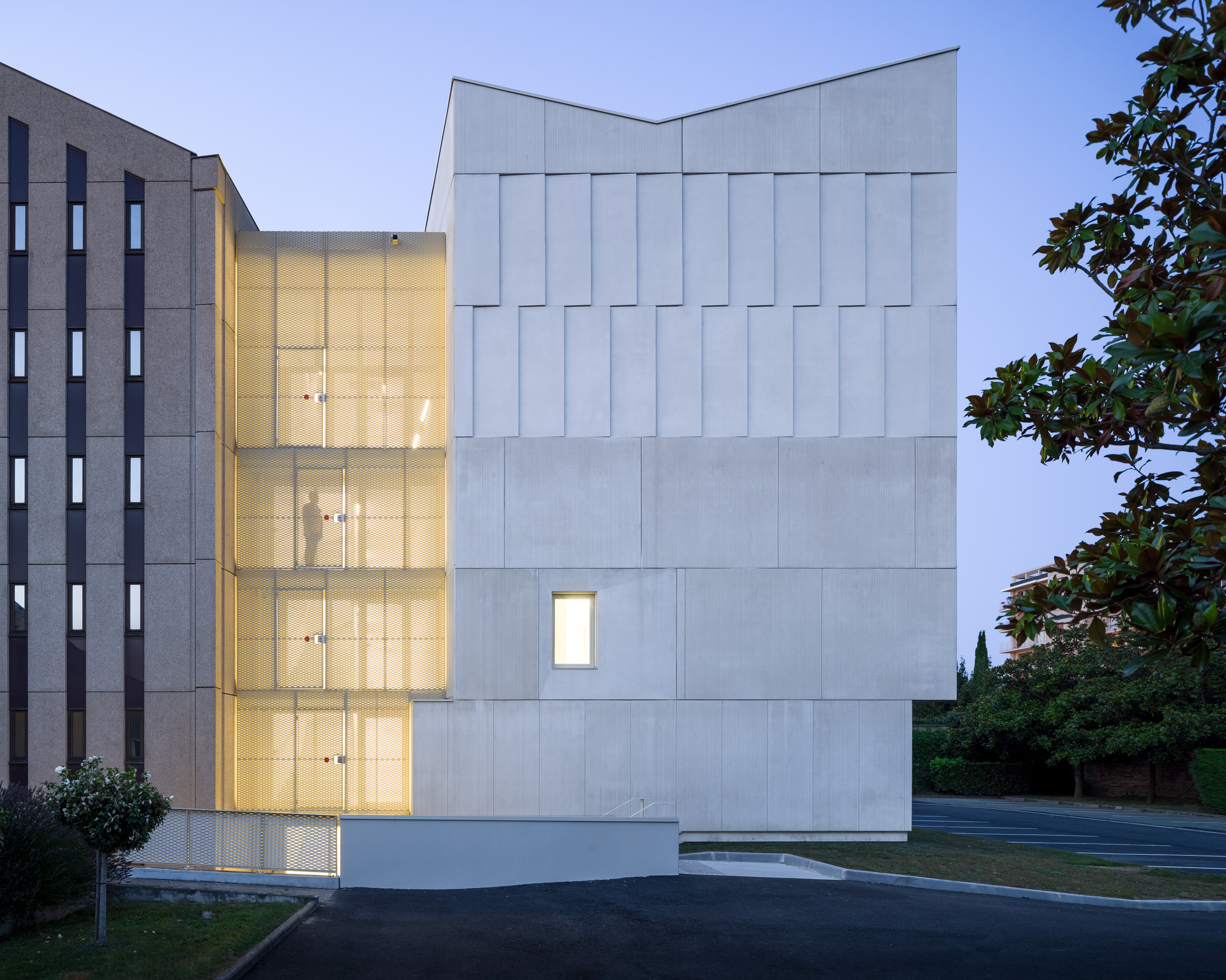 Dusk view of the white facade with illuminated glass stair tower connecting to existing concrete building