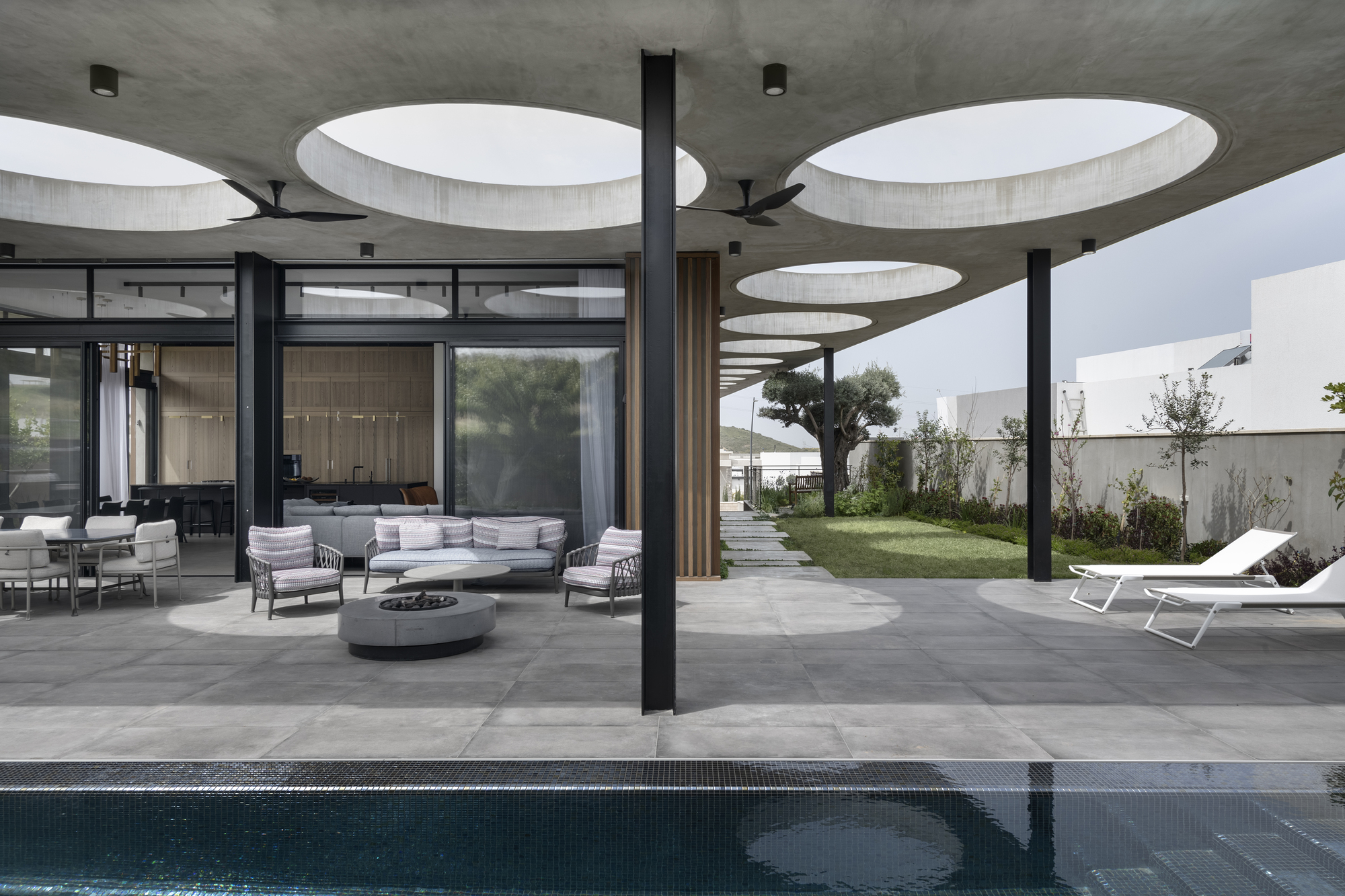 Covered outdoor terrace with circular ceiling voids and black columns overlooking a pool and garden