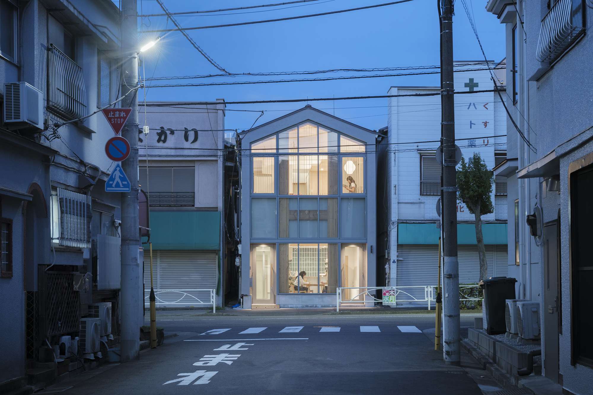 Gabled glass facade illuminated at twilight on a residential street with overhead power lines