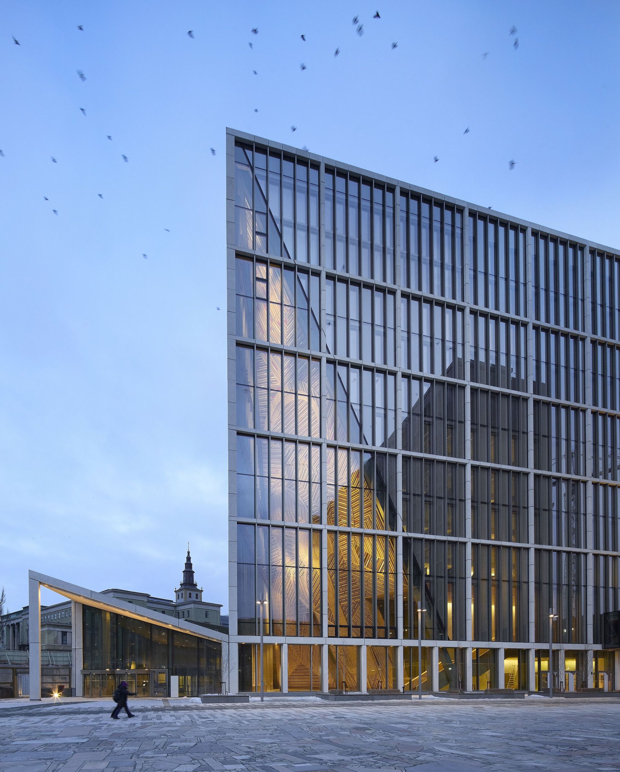 Glass facade reflecting the timber-clad atrium and surrounding buildings at dusk with birds overhead