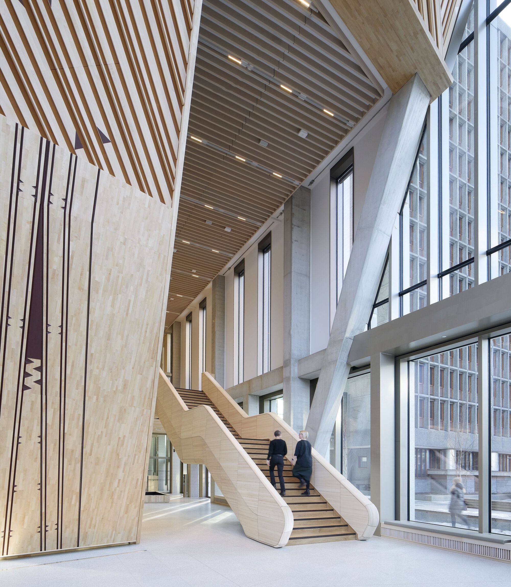 Light wood staircase ascending beneath angled structural beams and slatted ceiling with two people climbing