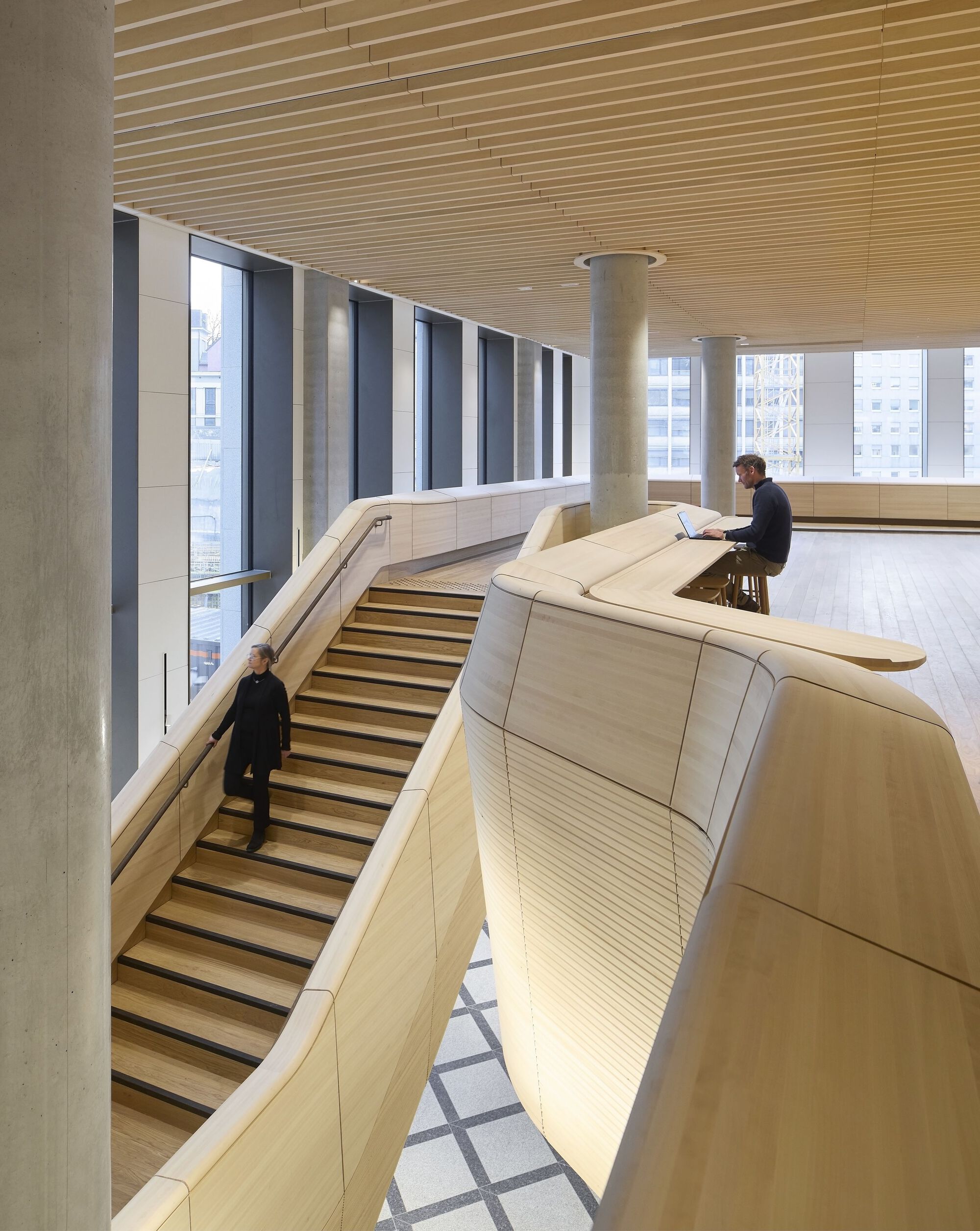 Curved bleacher seating with wood slat cladding beneath a linear wood batten ceiling as person works