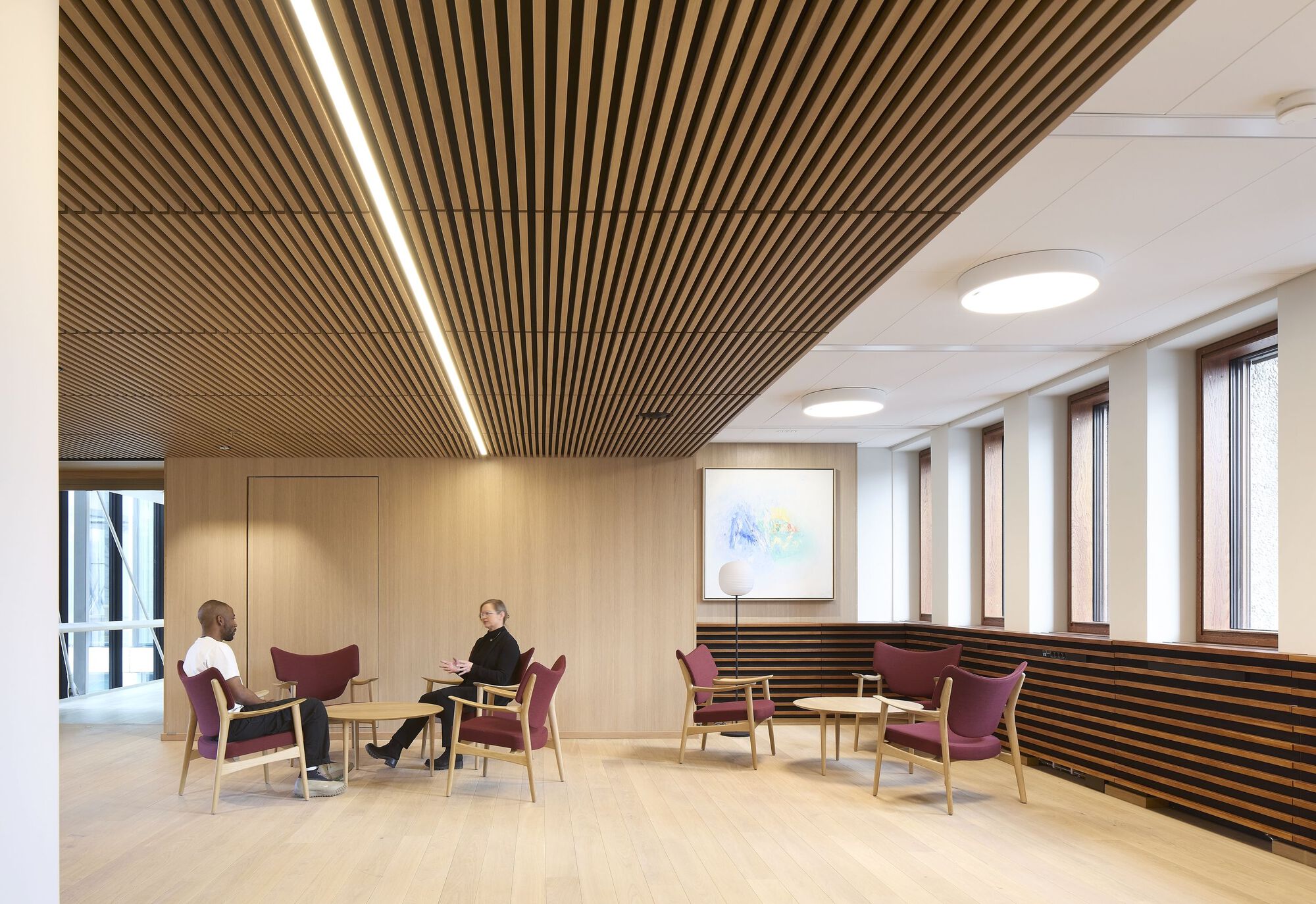 Meeting lounge with burgundy chairs beneath wood batten ceiling and circular skylights by windowed wall