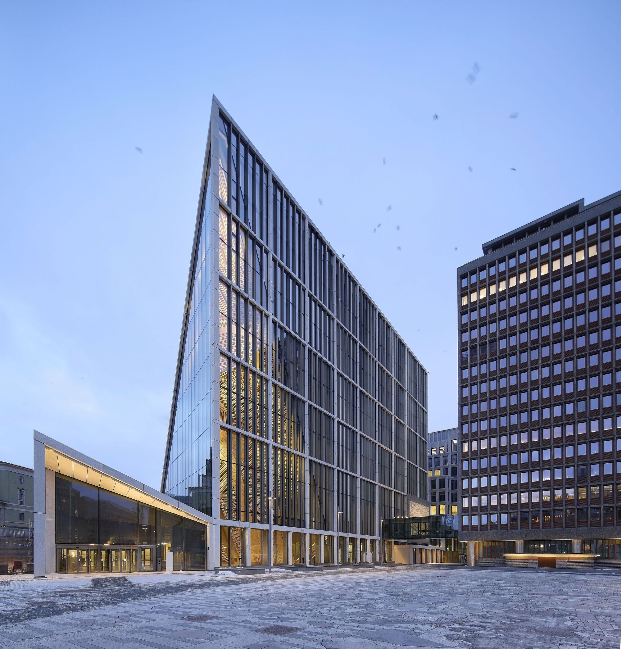 Angled glass and metal tower beside low glazed wing across paved plaza at dusk with birds overhead