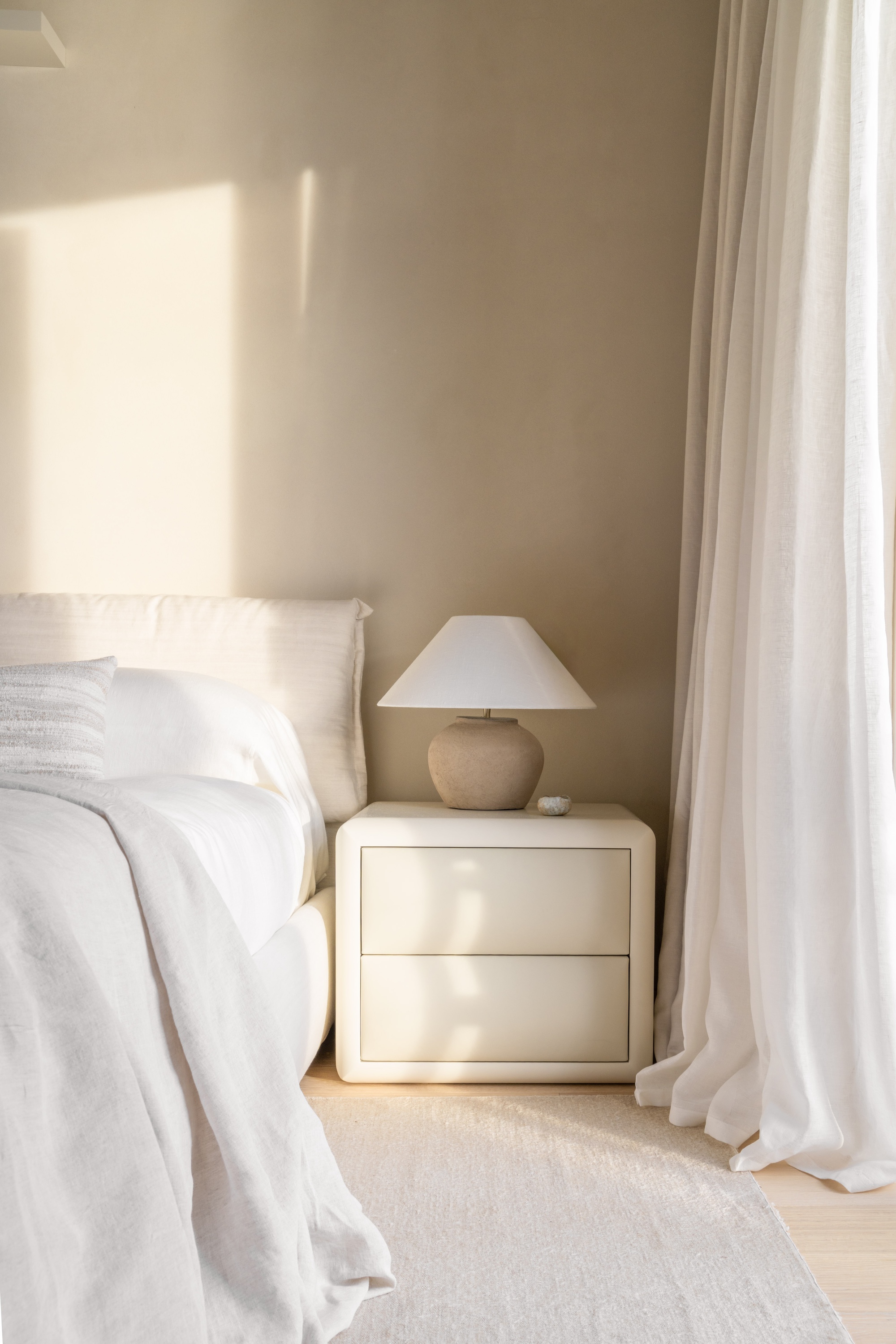 Bedside nightstand with table lamp beside white linen and floor-length curtains in soft morning light