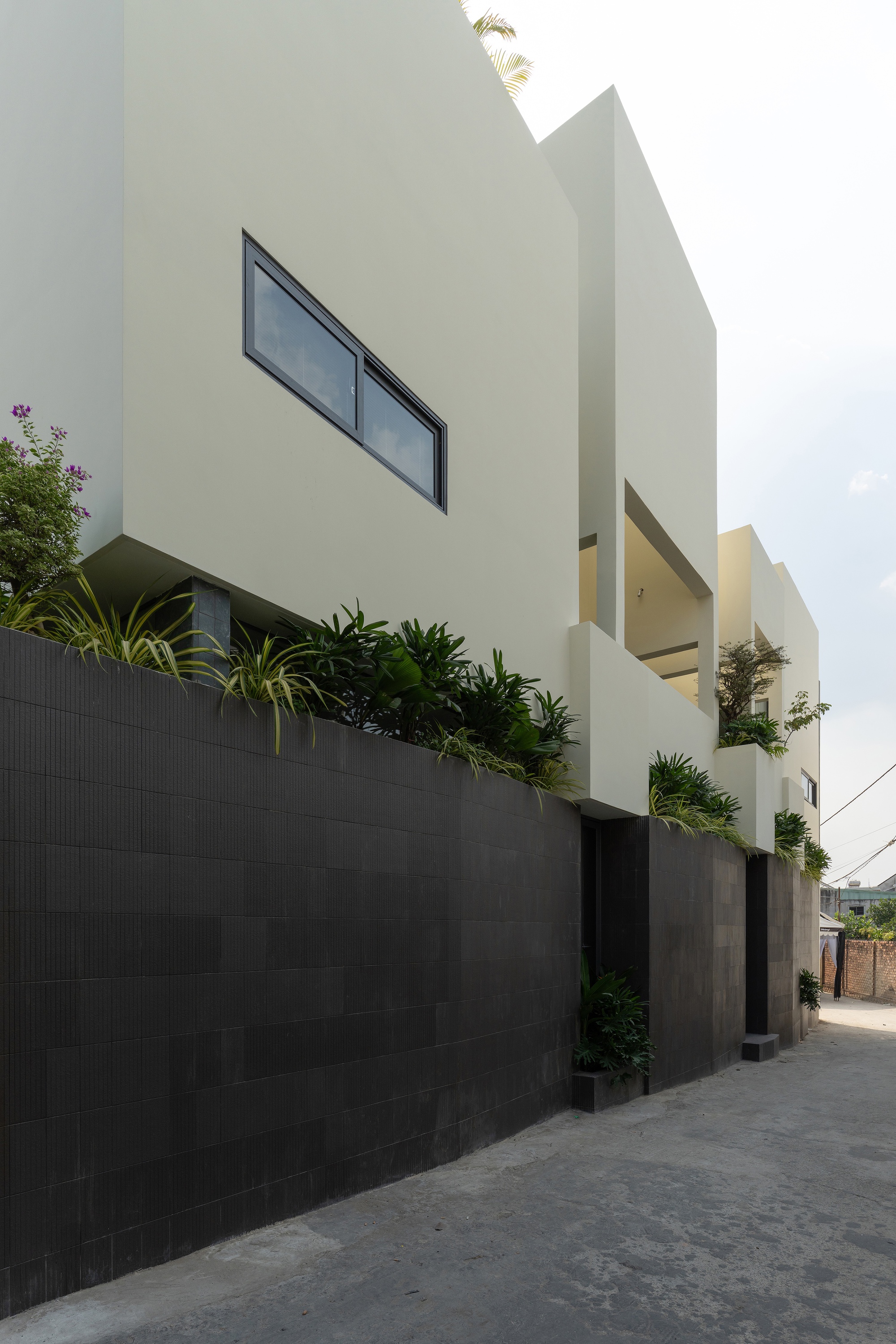 Street facade showing white geometric volumes rising above a dark tiled boundary wall with planted beds
