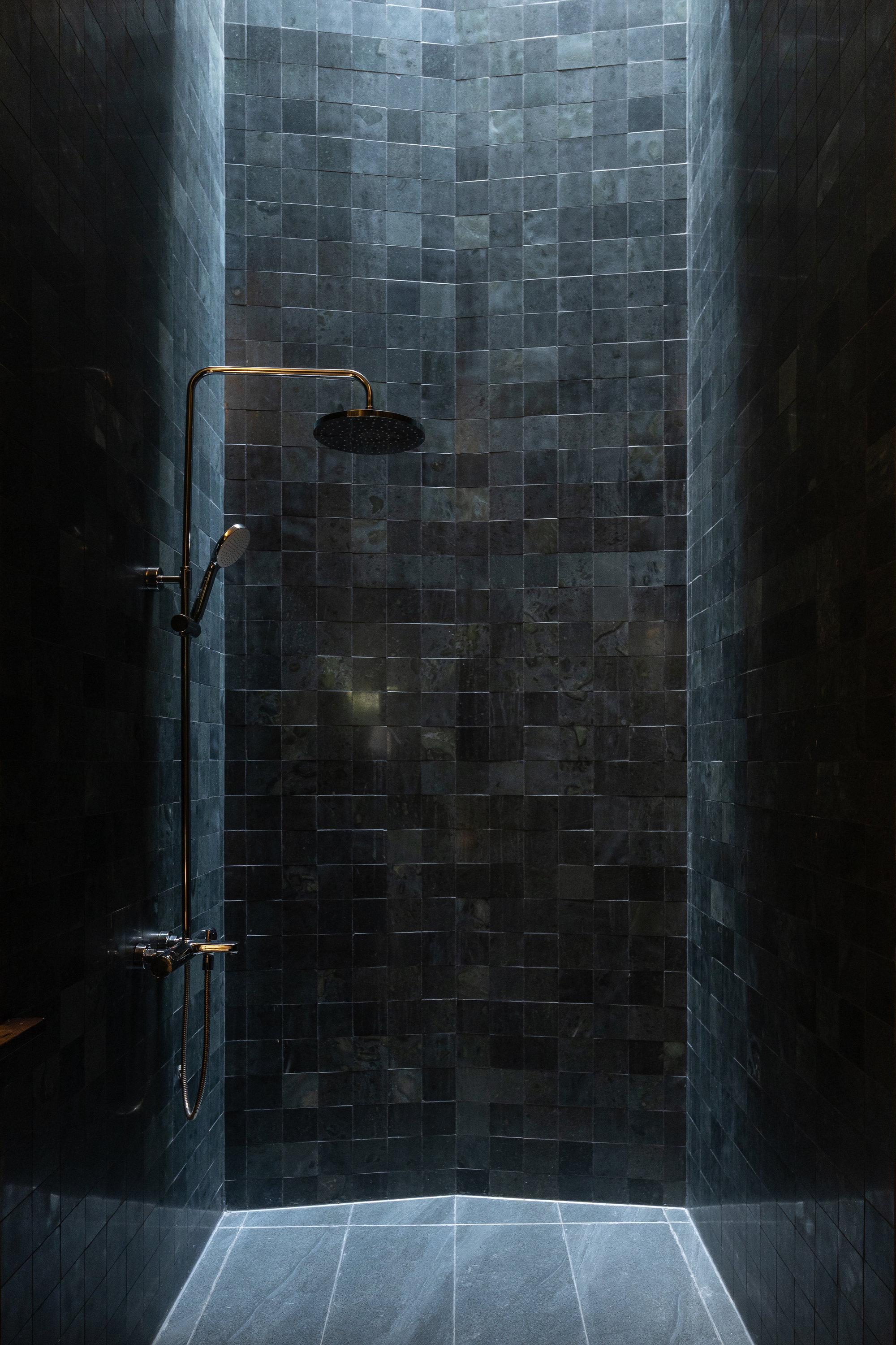 Shower enclosure with blue-grey glazed tile wall, brass fixtures, and backlit floor slit