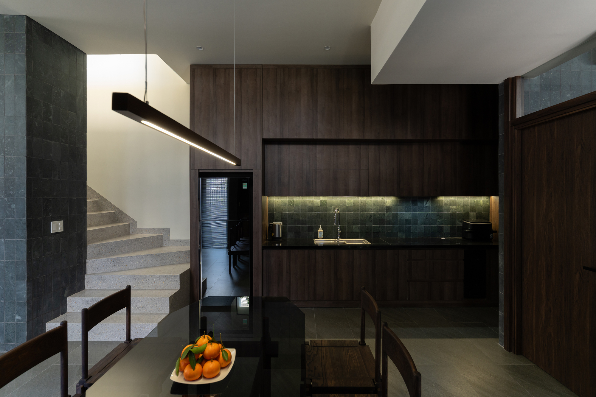 Open kitchen with dark wood cabinetry and under-cabinet lighting beside a concrete staircase with a fruit bowl