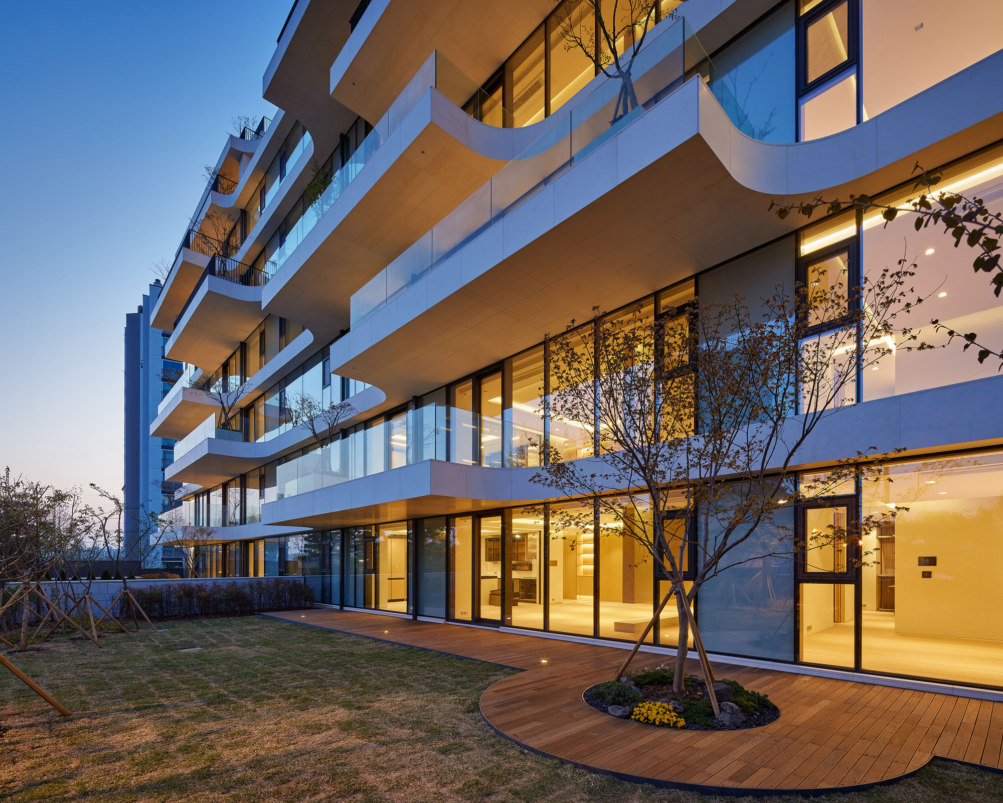 Stacked terraces with floor-to-ceiling glazing, planted trees and timber deck at twilight