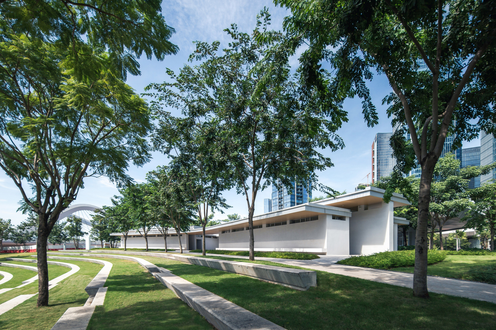Low-slung roof volume extending through a park with curved pathways and mature canopy trees