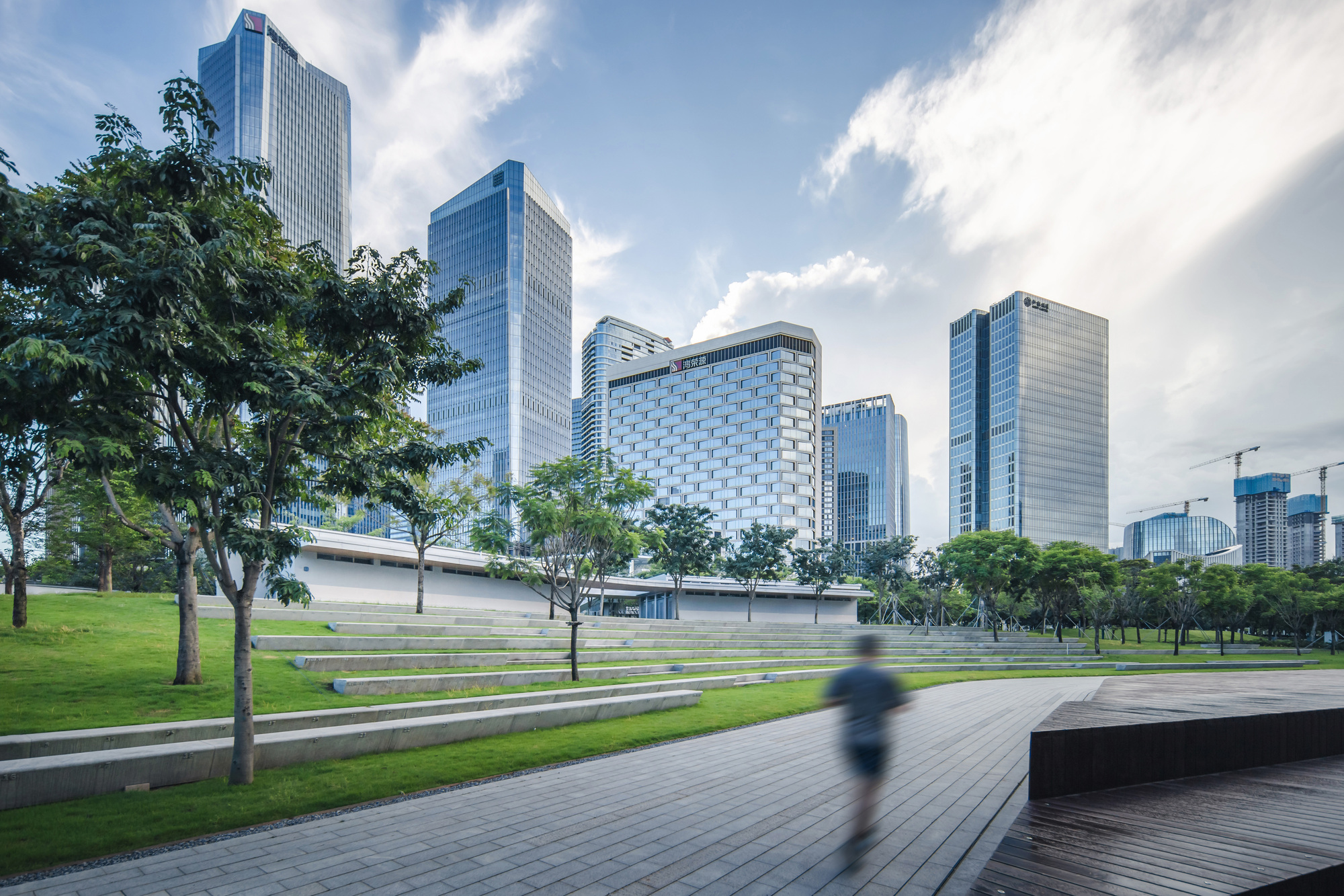 Curving paved plaza with terraced lawn and a pedestrian walking past scattered trees in daylight