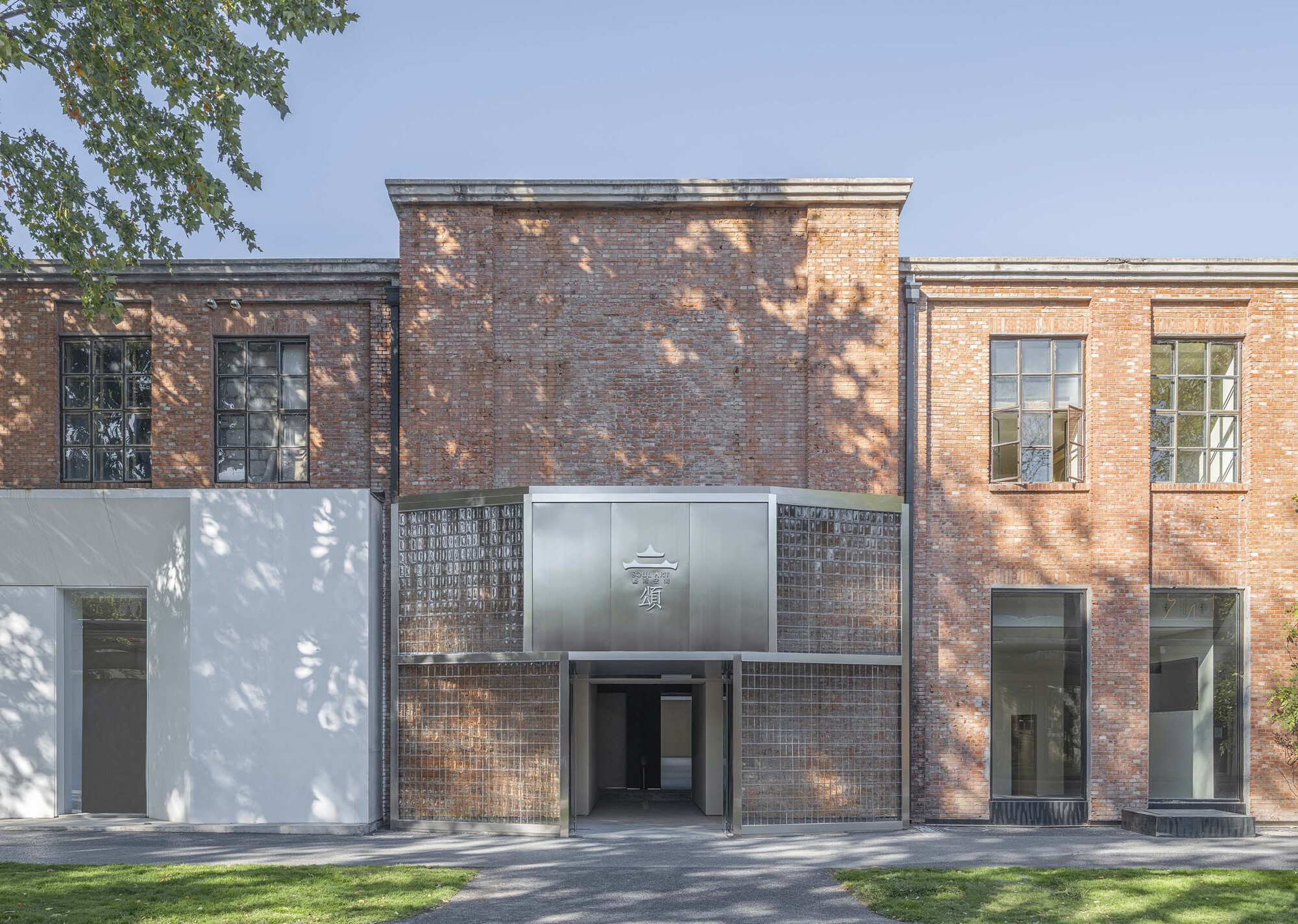 Front facade combining dark brick volumes with white stucco and glass brick under dappled tree shadow
