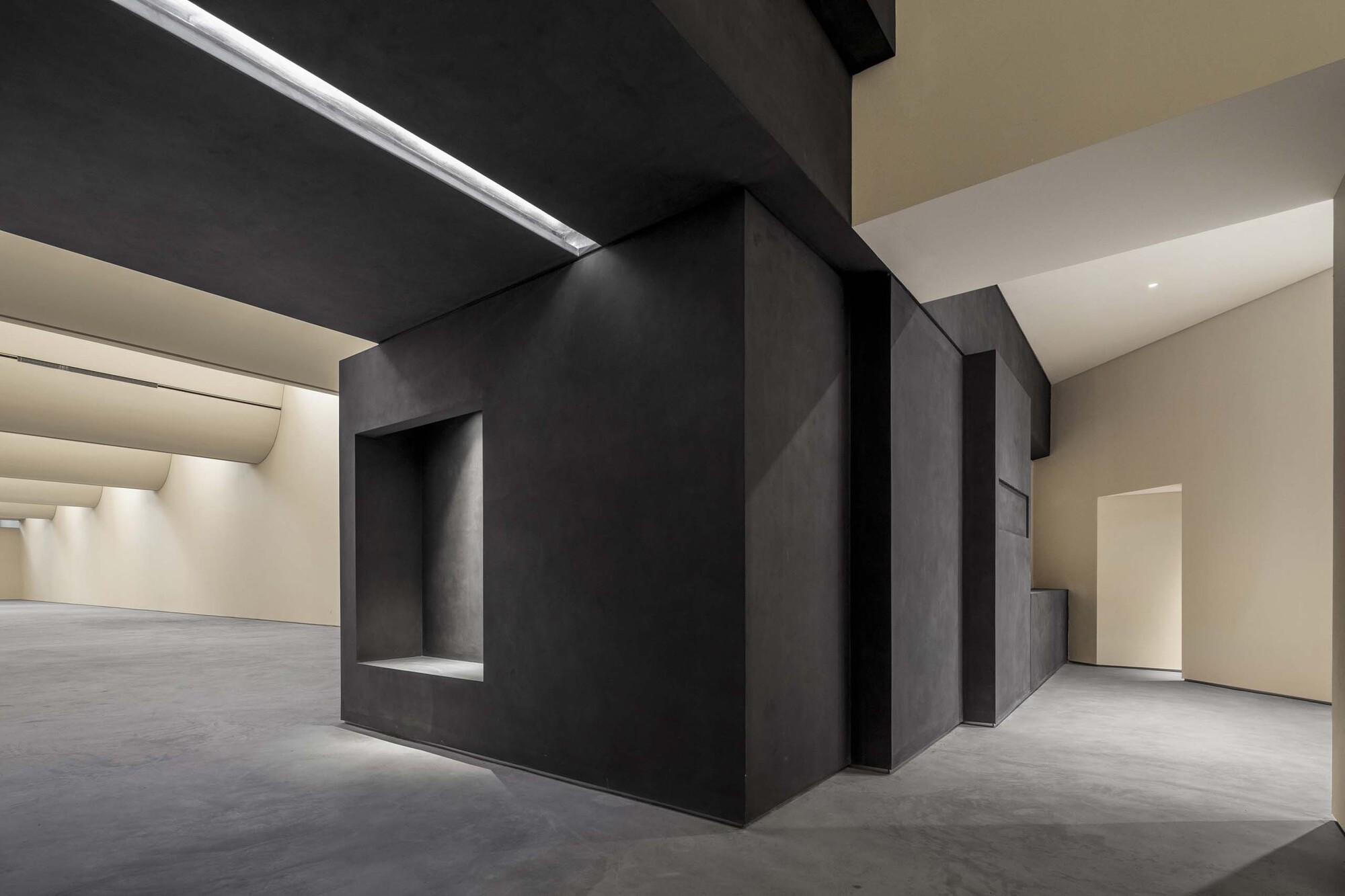 Dark metallic exhibition wall with recessed niche alongside white gallery spaces under artificial light