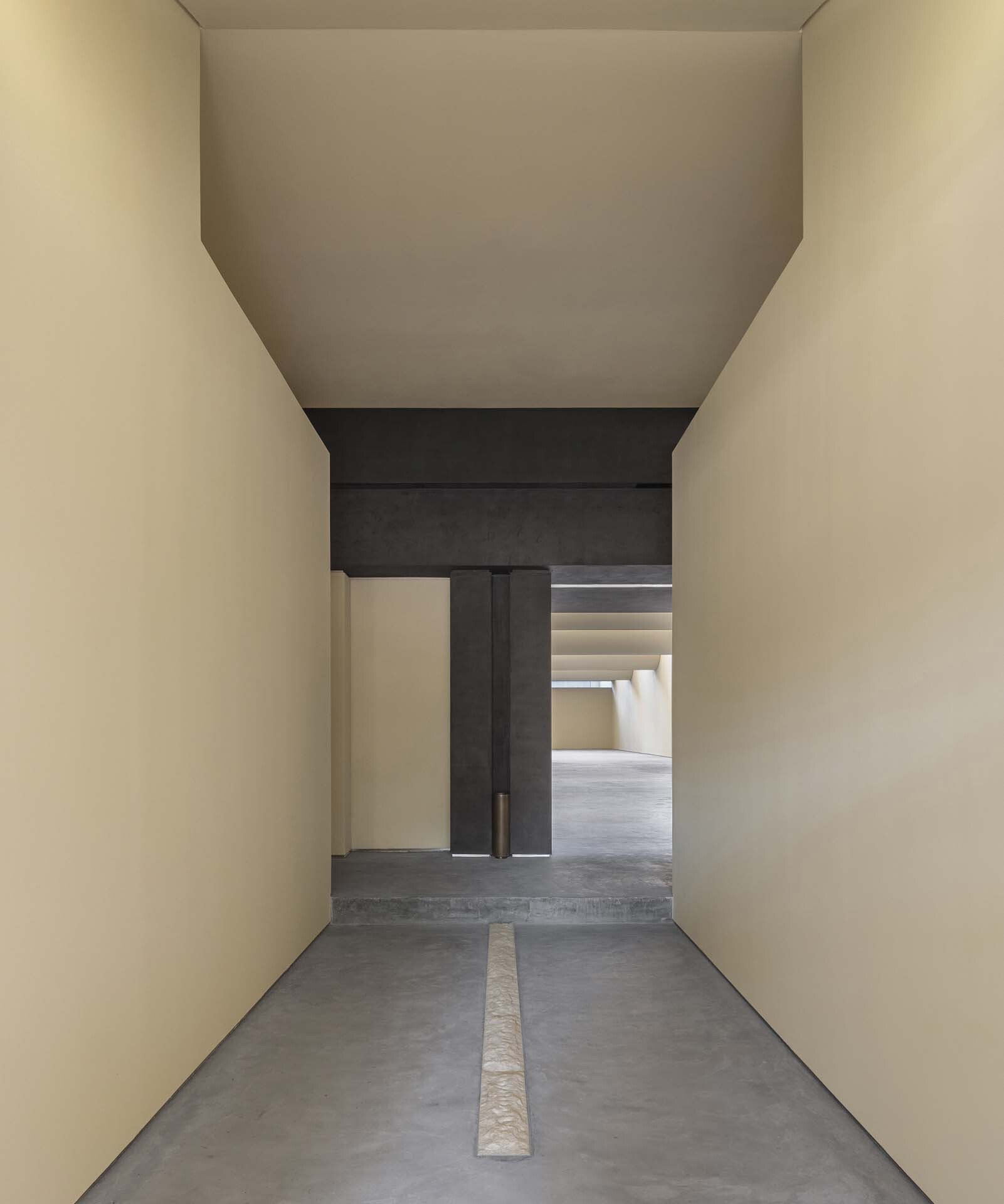 Narrow passage with limestone floor inlay leading through dark concrete frame into stepped ceiling gallery