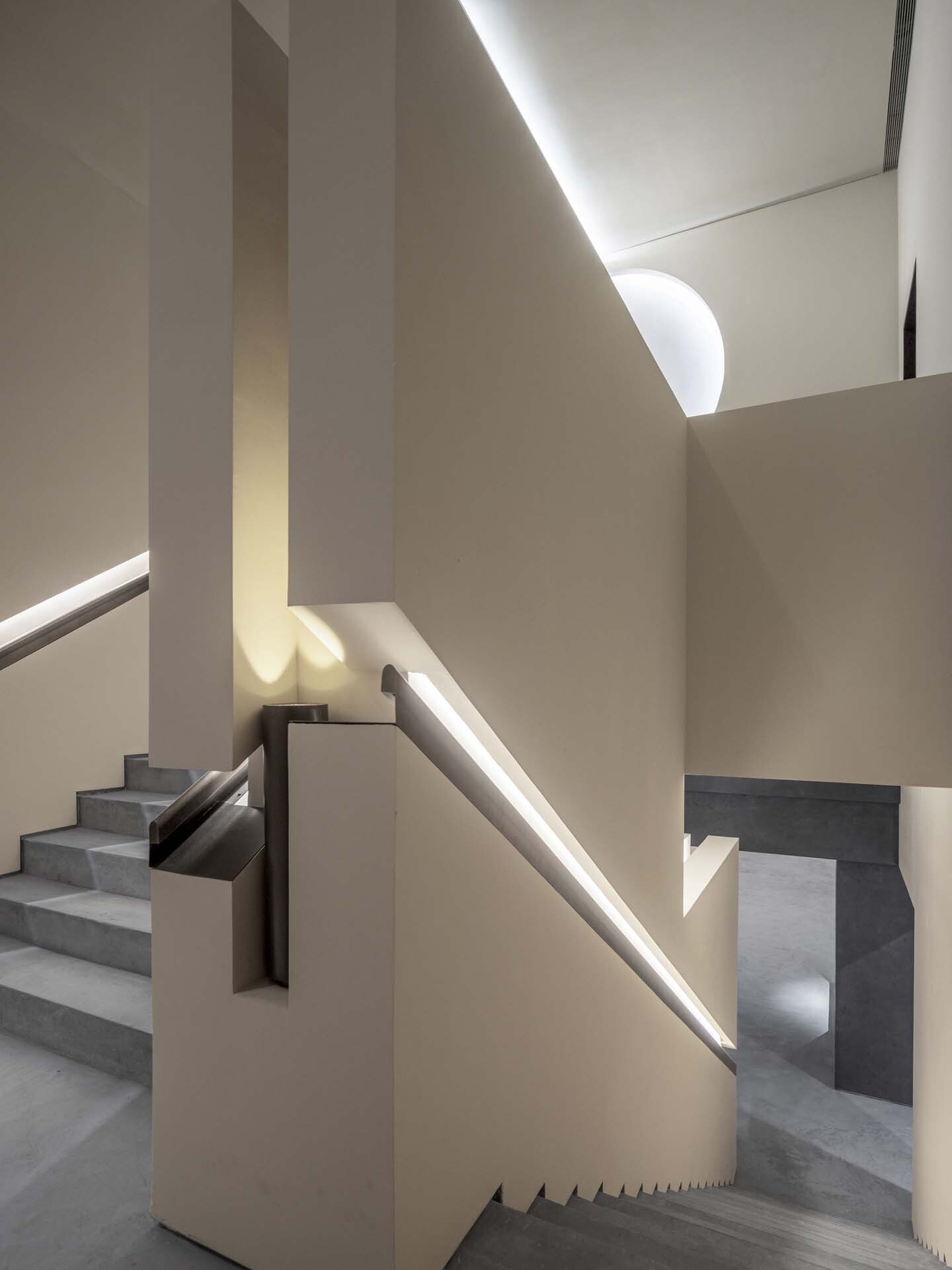 Concrete staircase with integrated LED handrail lighting ascending between cream plaster walls and skylights