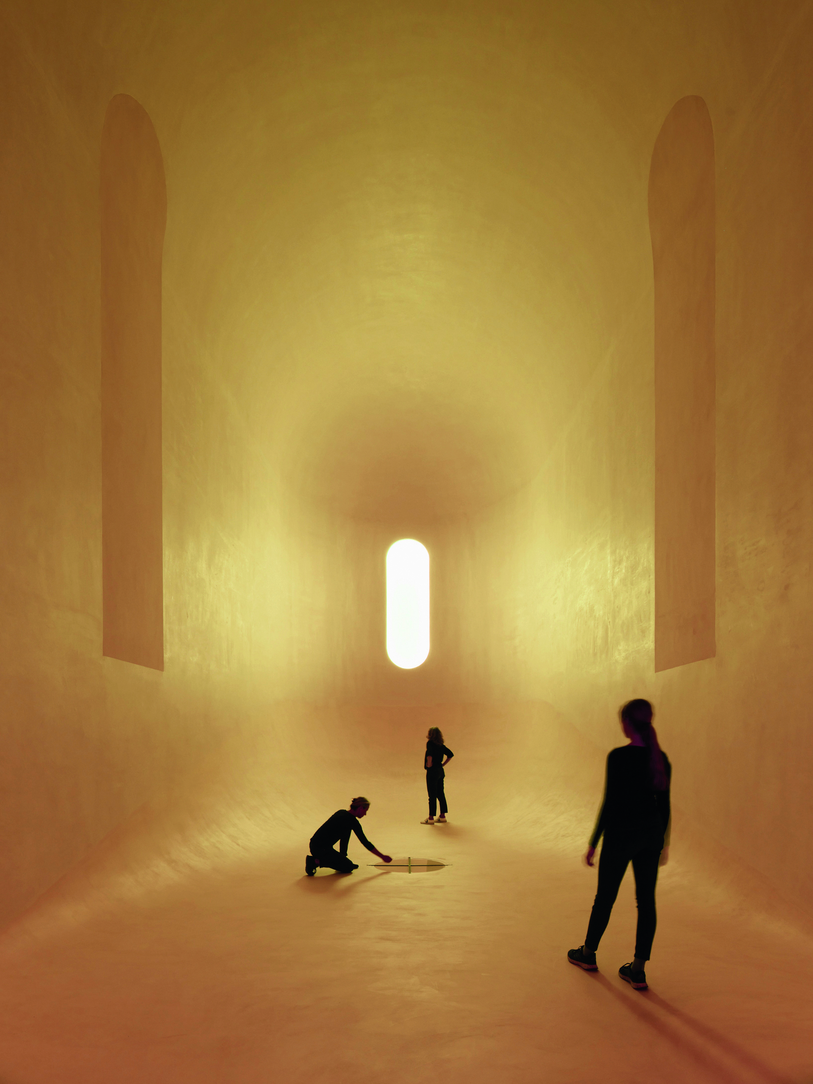 Golden-lit vaulted interior with tall vertical niches and visitors gathered beneath a single capsule-shaped window