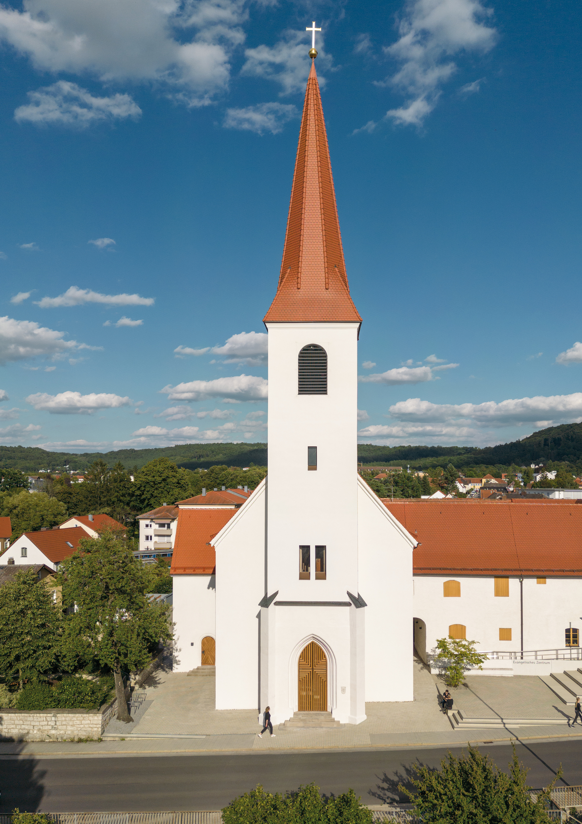 Front elevation of a white church with terracotta spire and timber arched door under a bright blue sky
