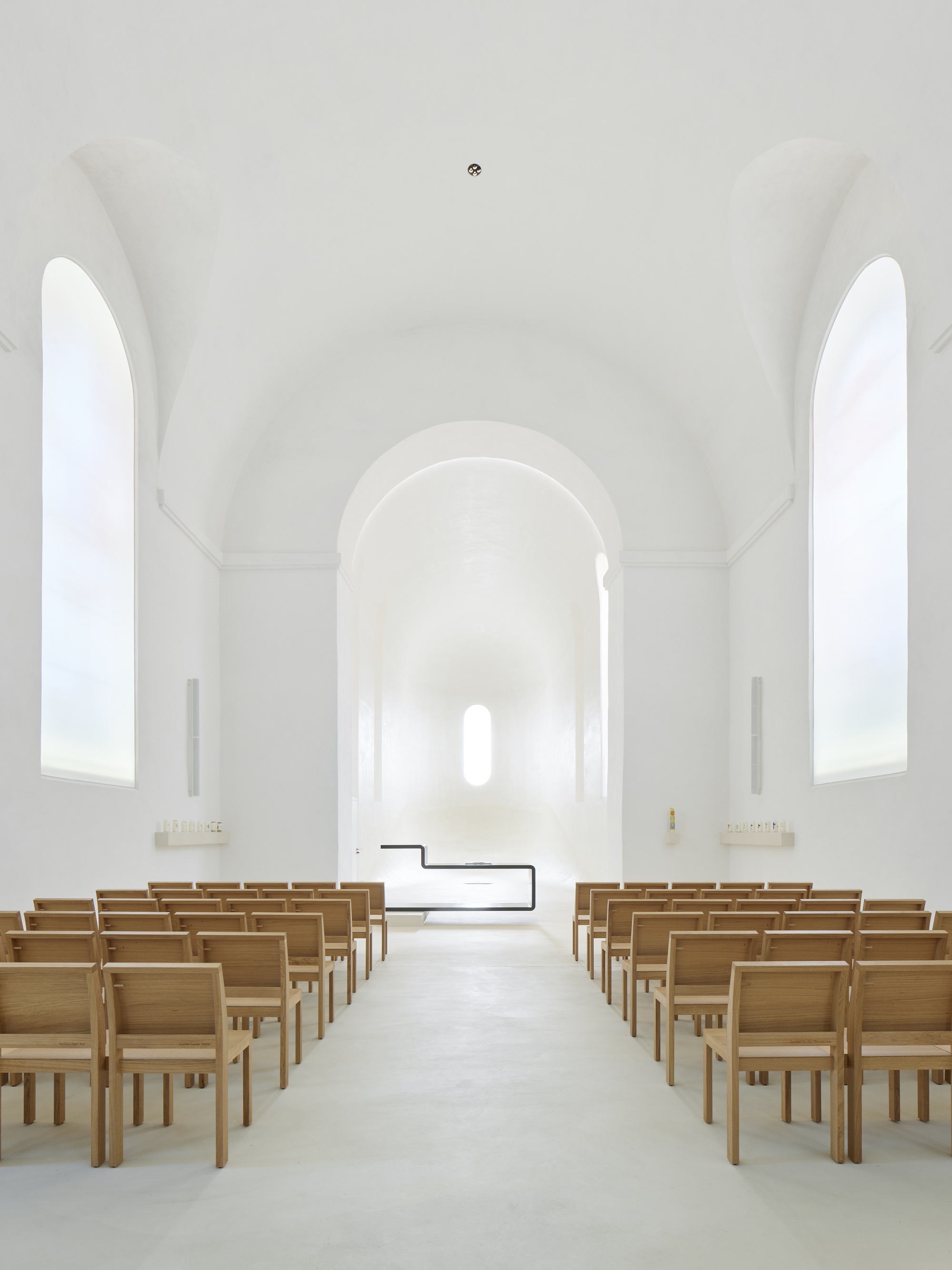White vaulted chapel interior with timber pews flanking a central aisle leading to arched apse
