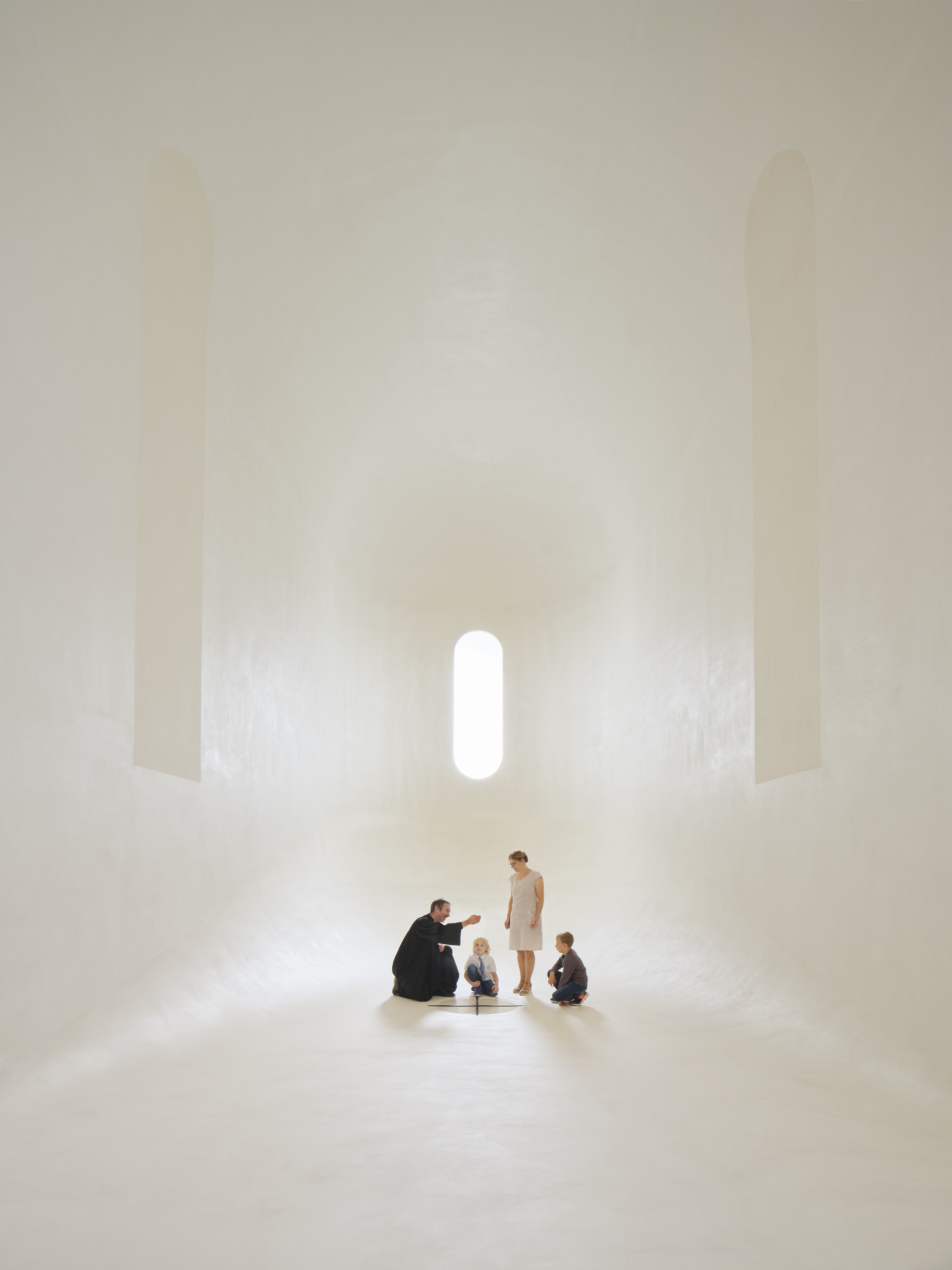 Apsidal sanctuary space with family group gathered on white terrazzo floor under vertical light slot