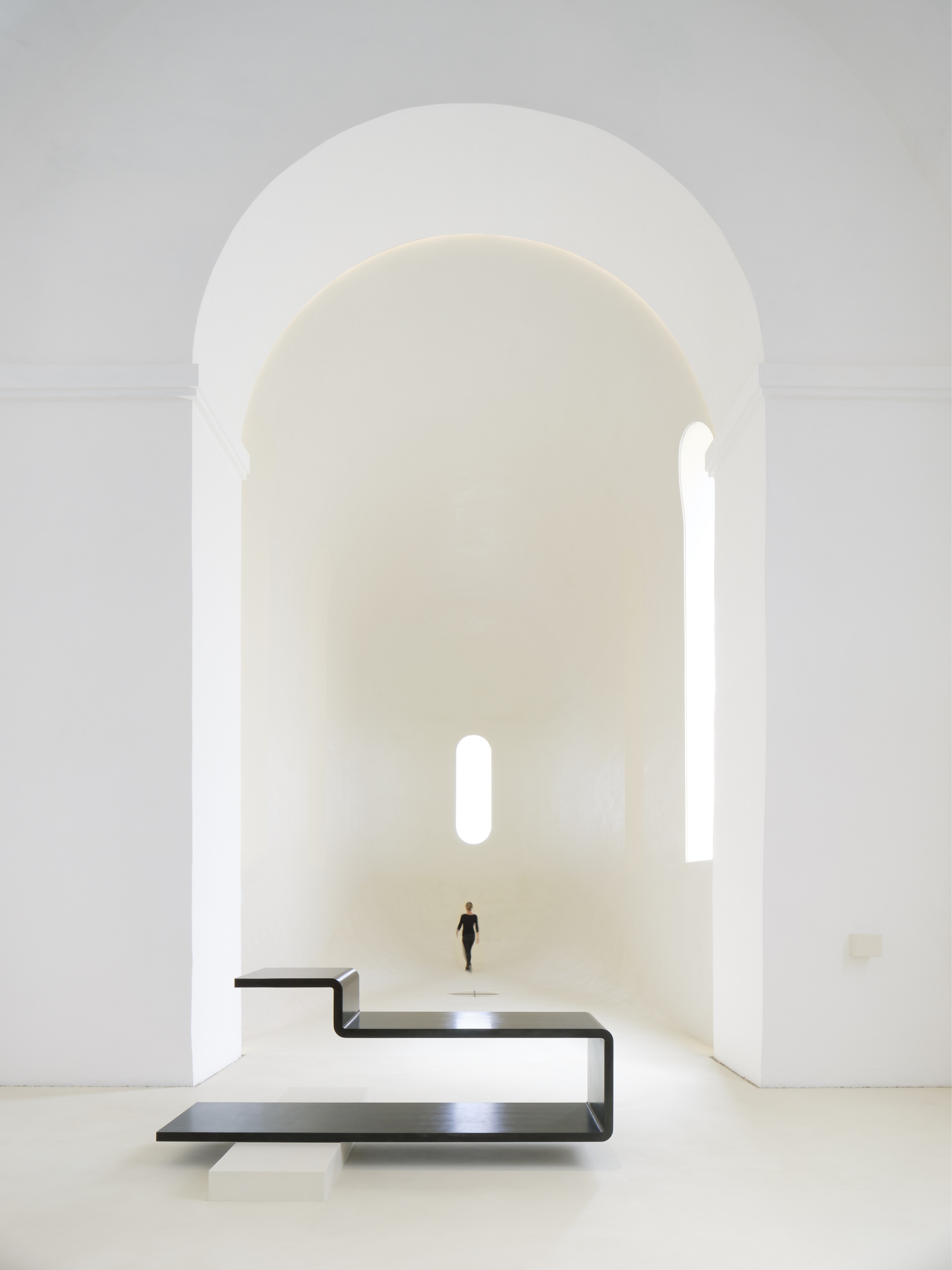 Cantilevered black steel platform in white nave with lone figure walking toward apse