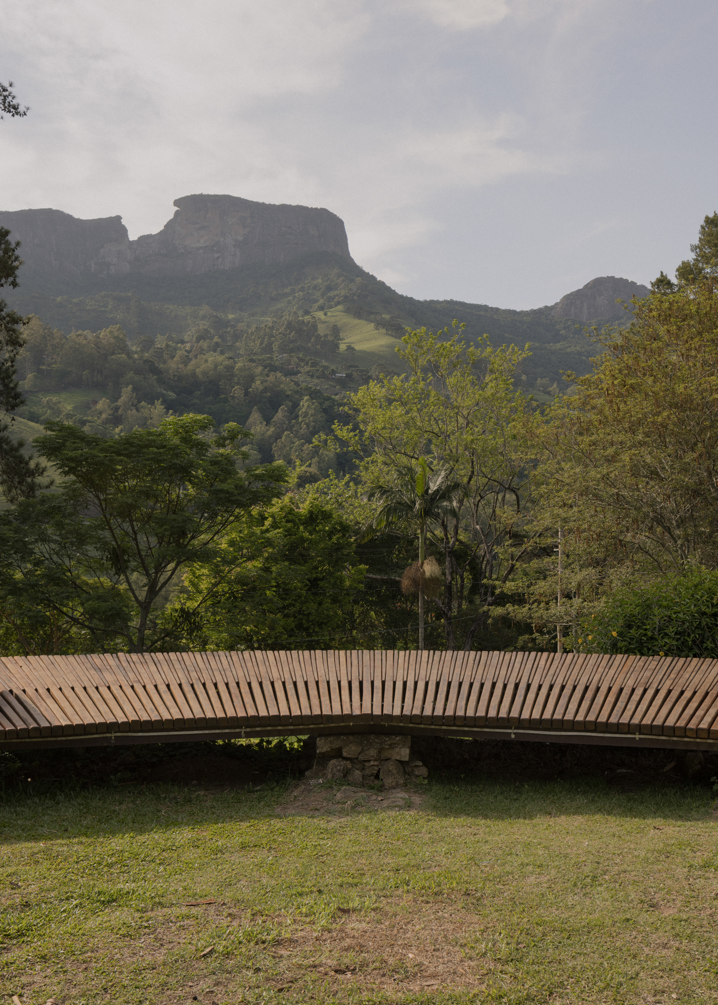 Horizontal slatted timber bridge spanning grass with mountain cliffs rising behind in mist